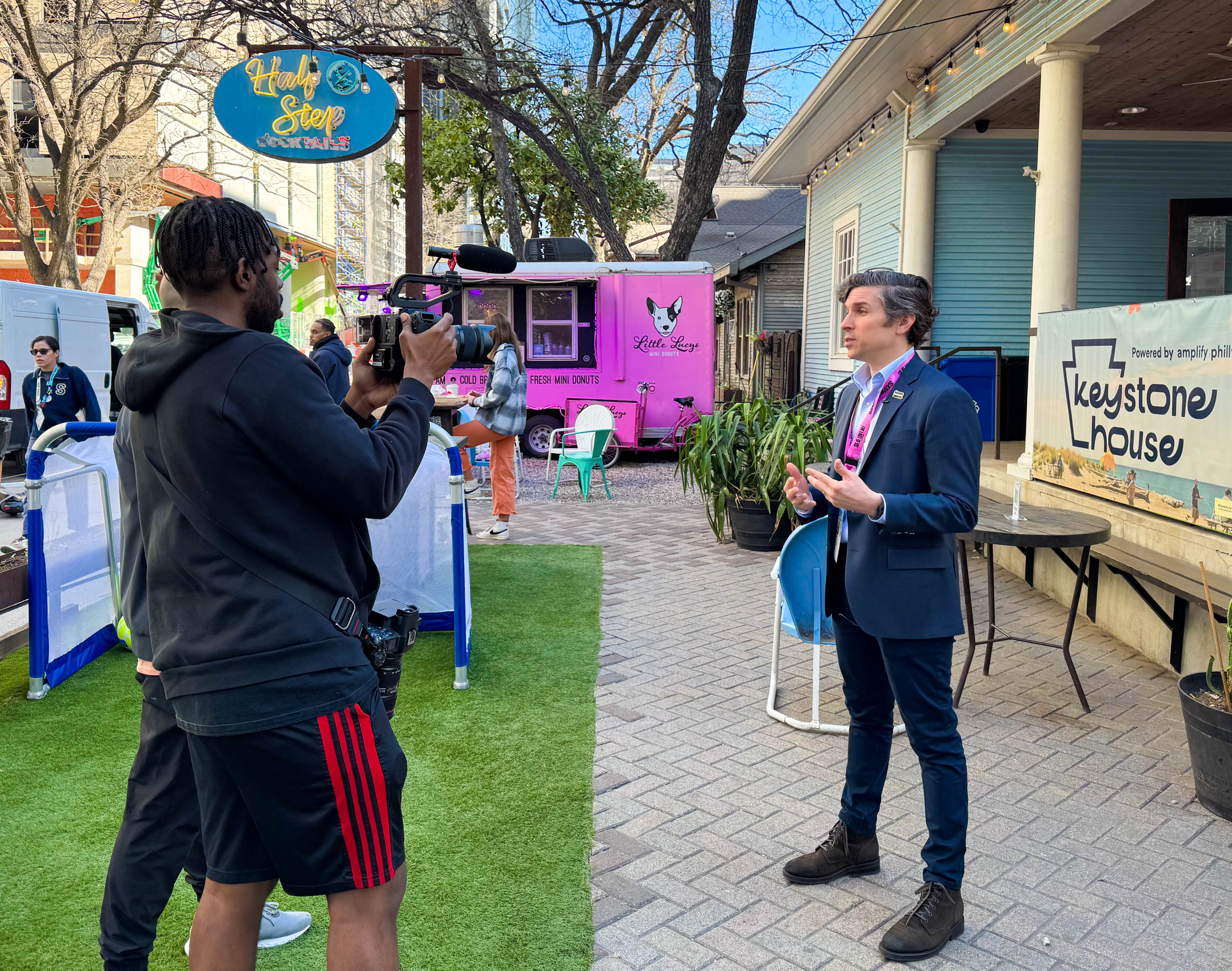 Michael O'Rourke is interviewed by two videographers at an outdoor event at SXSW. A food truck and crowd are visible in the distance.
