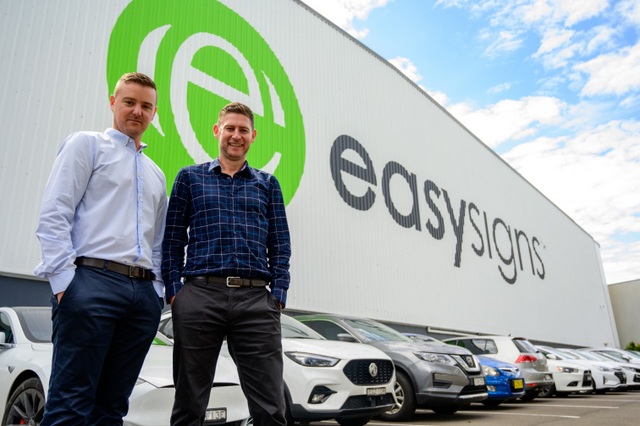 Two men in business attire smile for a photo, standing in front of a large warehouse. The warehouse has a logo for the company Easy Signs on it, and cars are parked in front of the building.