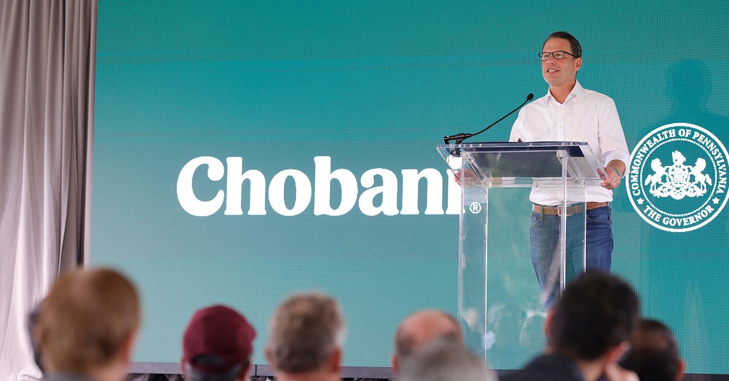 Governor Josh Shapiro speaks at a podium to a crowd of businesspeople. The Chobani logo is visible behind him.