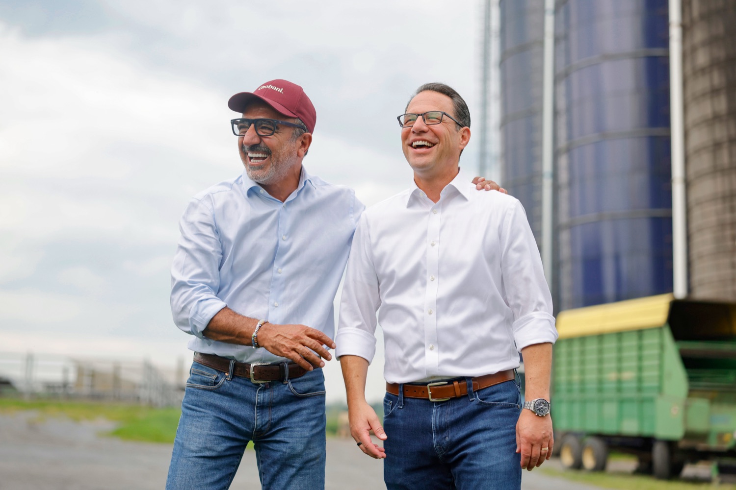 The CEO of Chobani and Pennsylvania Governor Josh Shapiro stand alongside each other and smile. A farm silo and field are in the background..