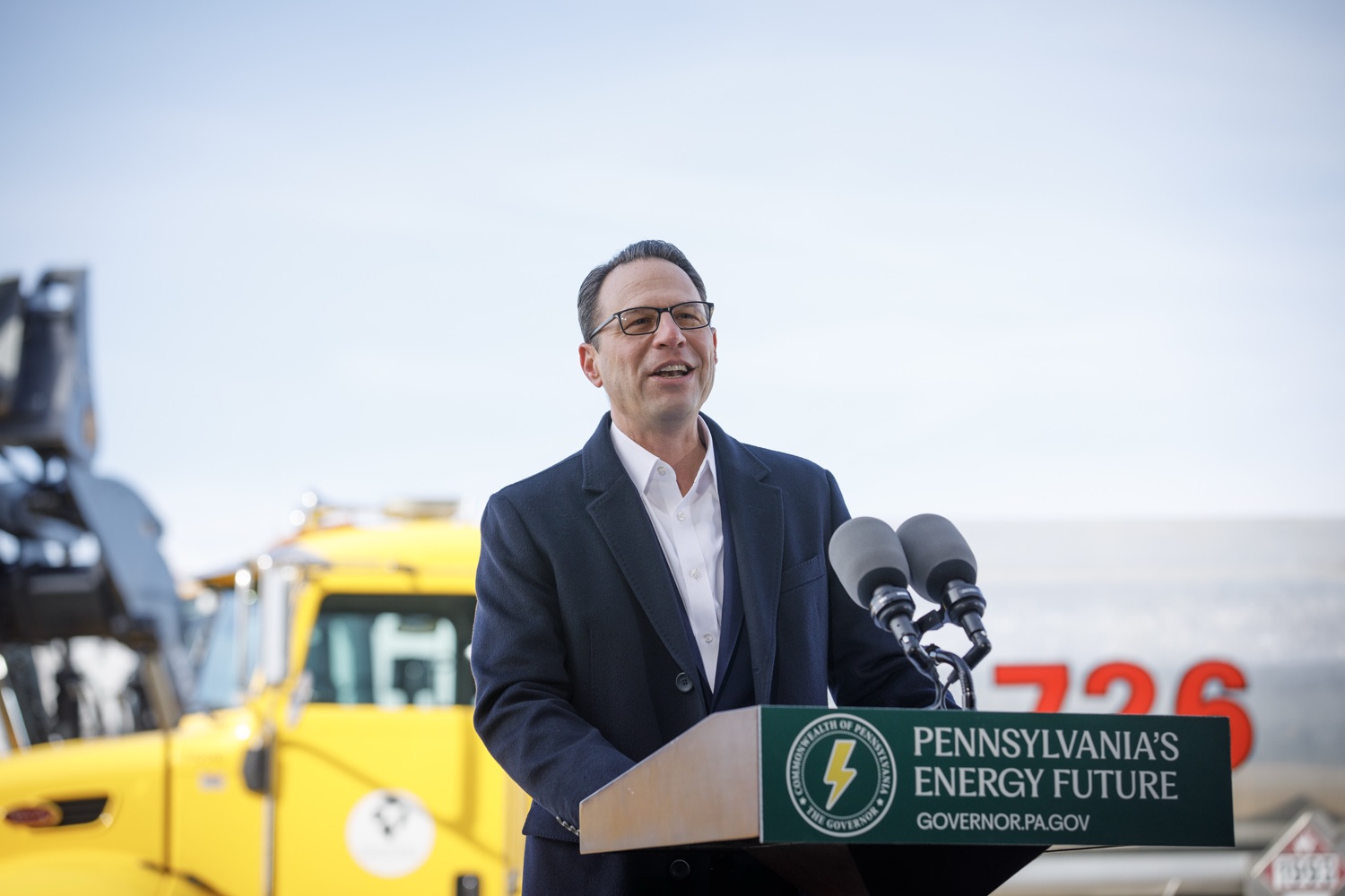 Governor Josh Shapiro, standing at a podium on the tarmac of Pittsburgh International Airport, announces an energy plan. Construction vehicles are visible in the background.