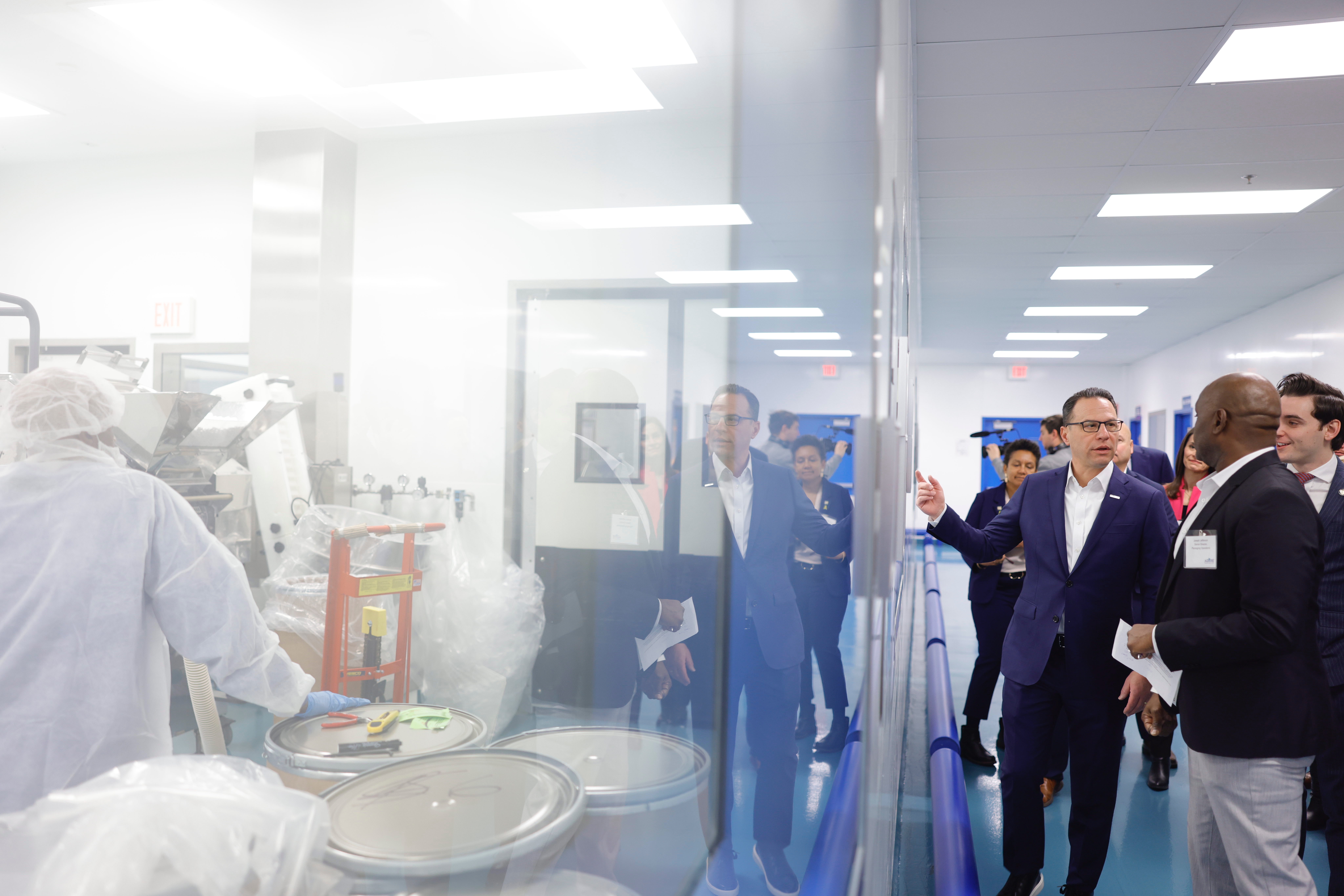 Gov. Josh Shapiro and executives tour a biopharma facility, viewing lab workers working through a large glass window.