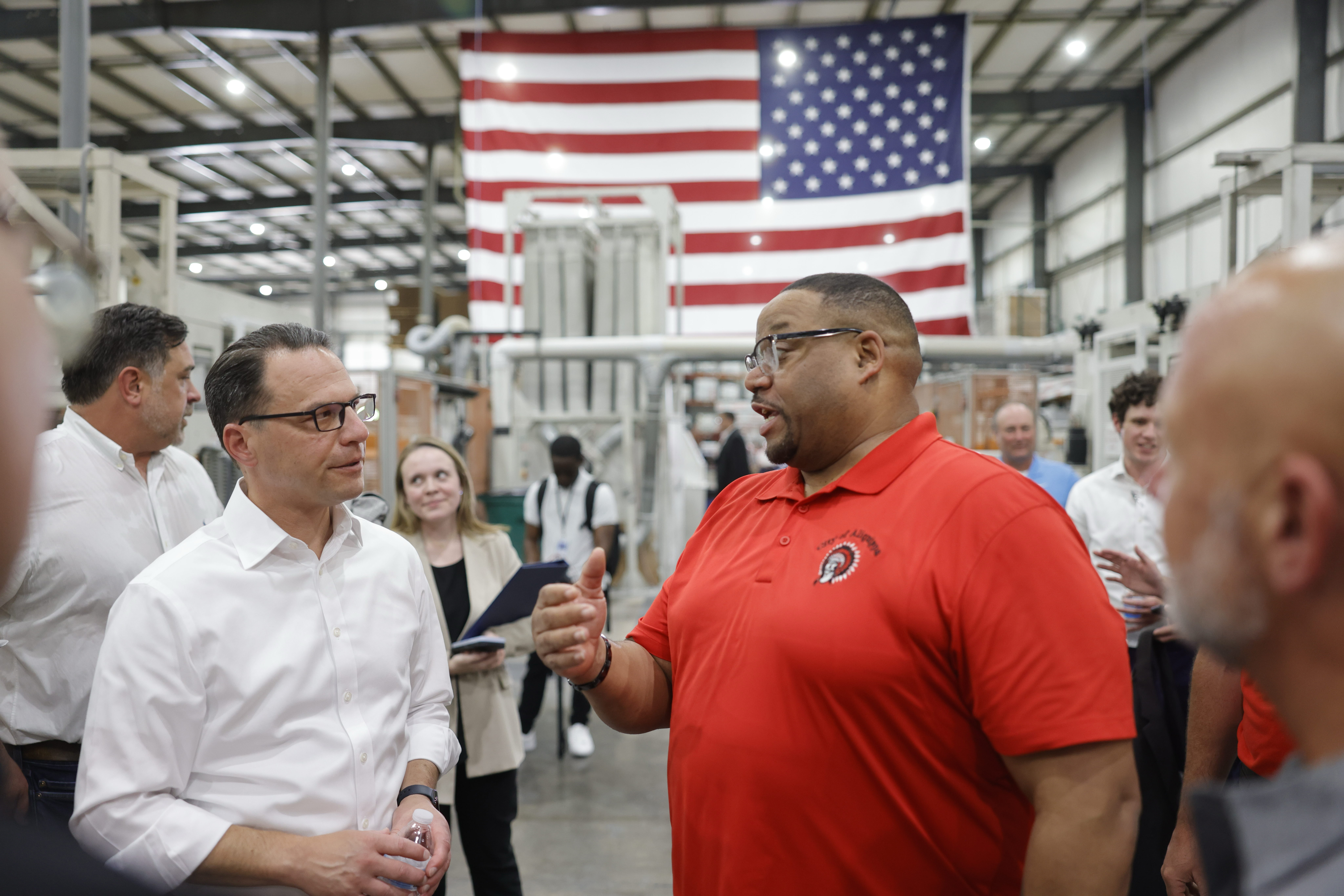 Gov. Josh Shapiro talks with a Versatex employee in a tour of the manufacturing floor. A large American flag hangs from the ceiling over a small crowd of executives.