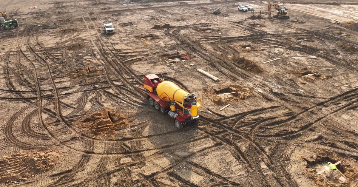 A cement mixer drives across a dirt construction lot.