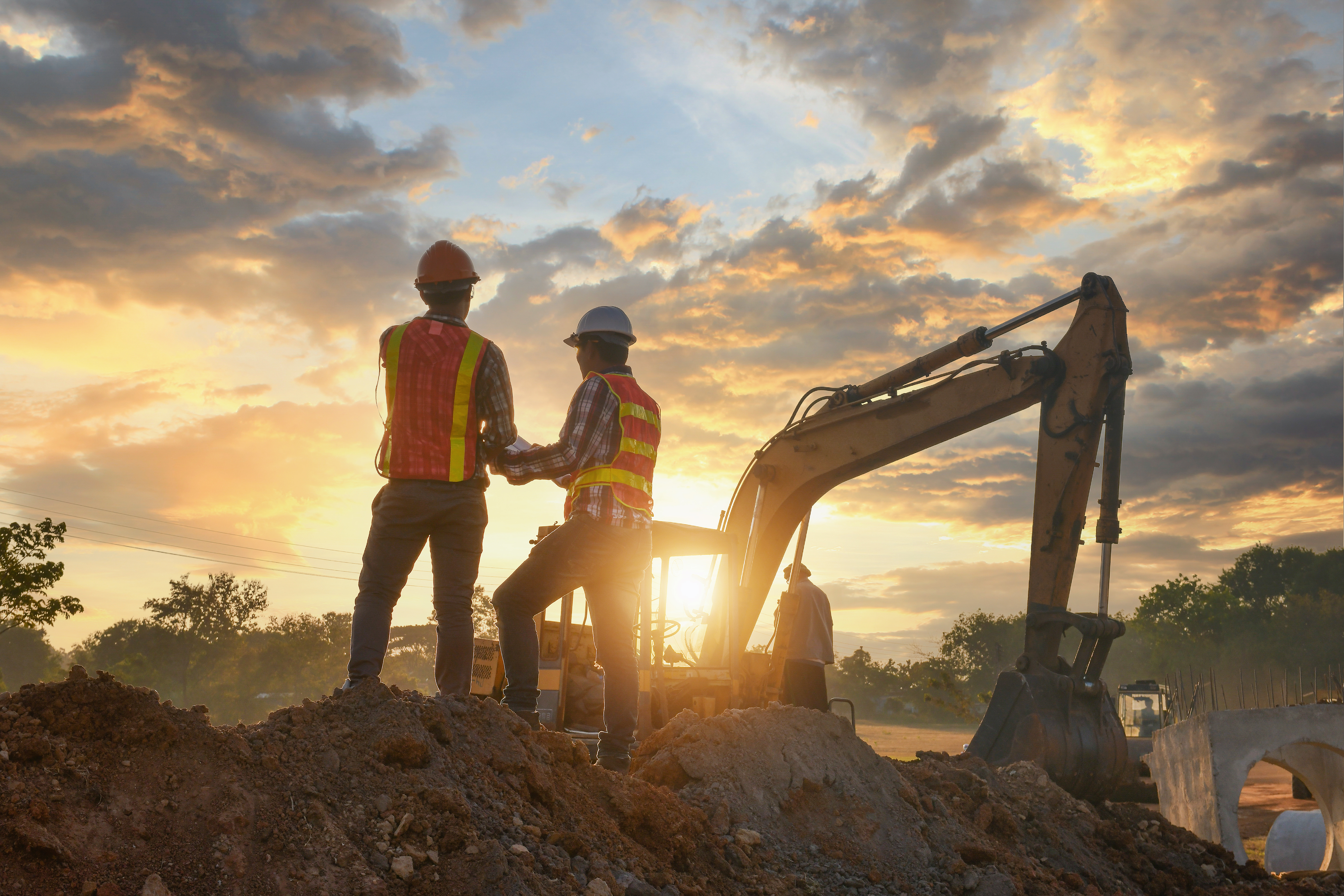 Two silohuetted construction workers talk in front of an excavator at a construction site during sunset