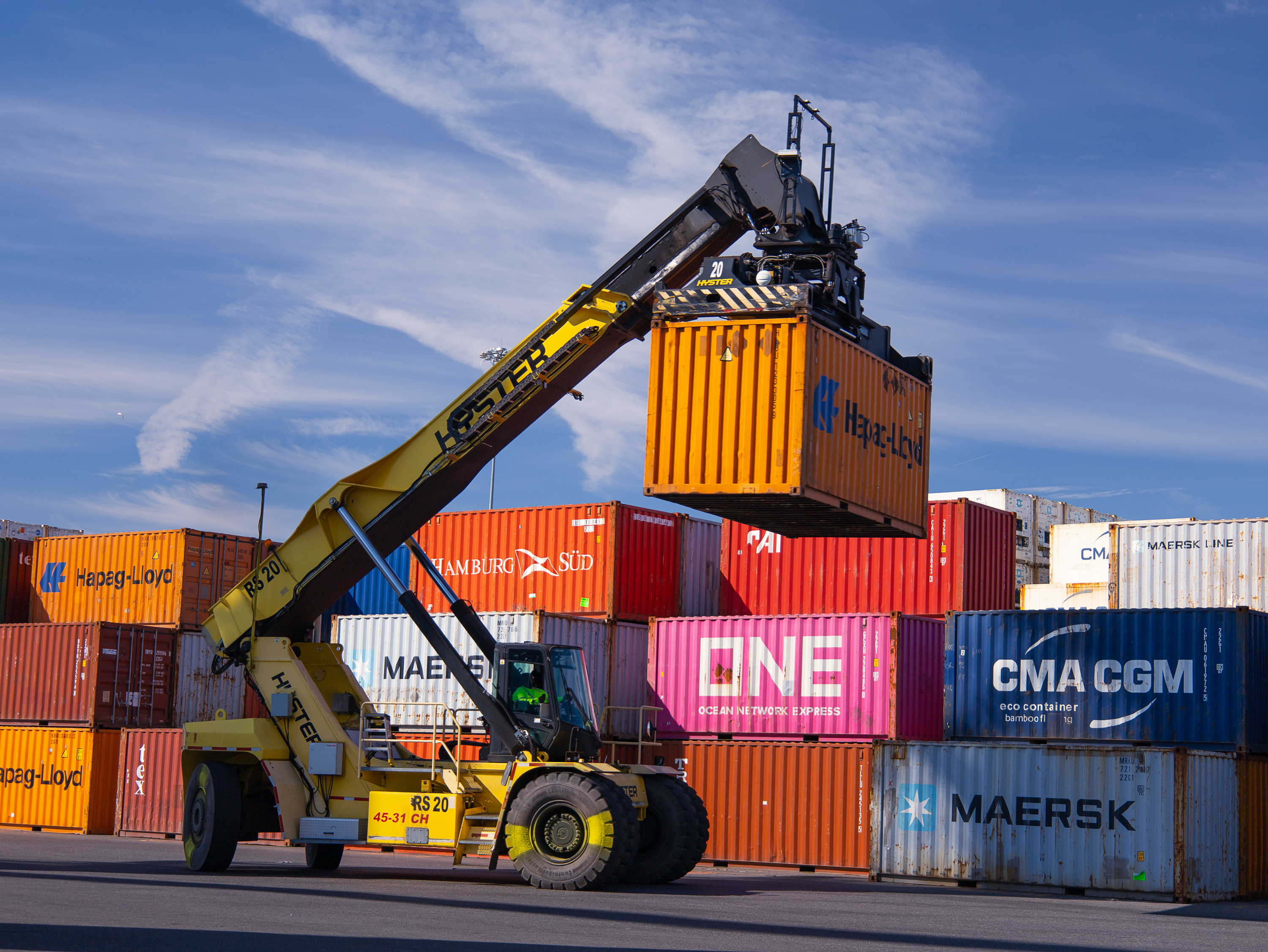 A mobile crane lifts a shipping container onto a stack of colorful shipping containers at a port