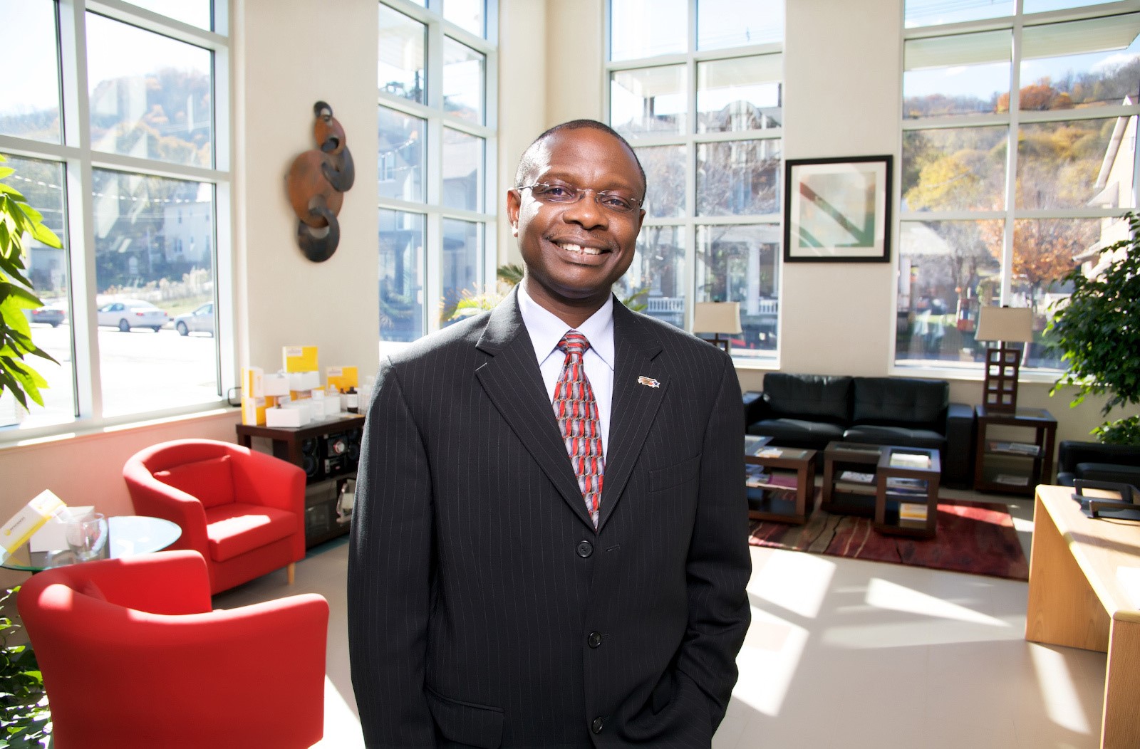 A man wearing a suit smiles for a picture in a brightly lit office lobby with many windows and modern furniture
