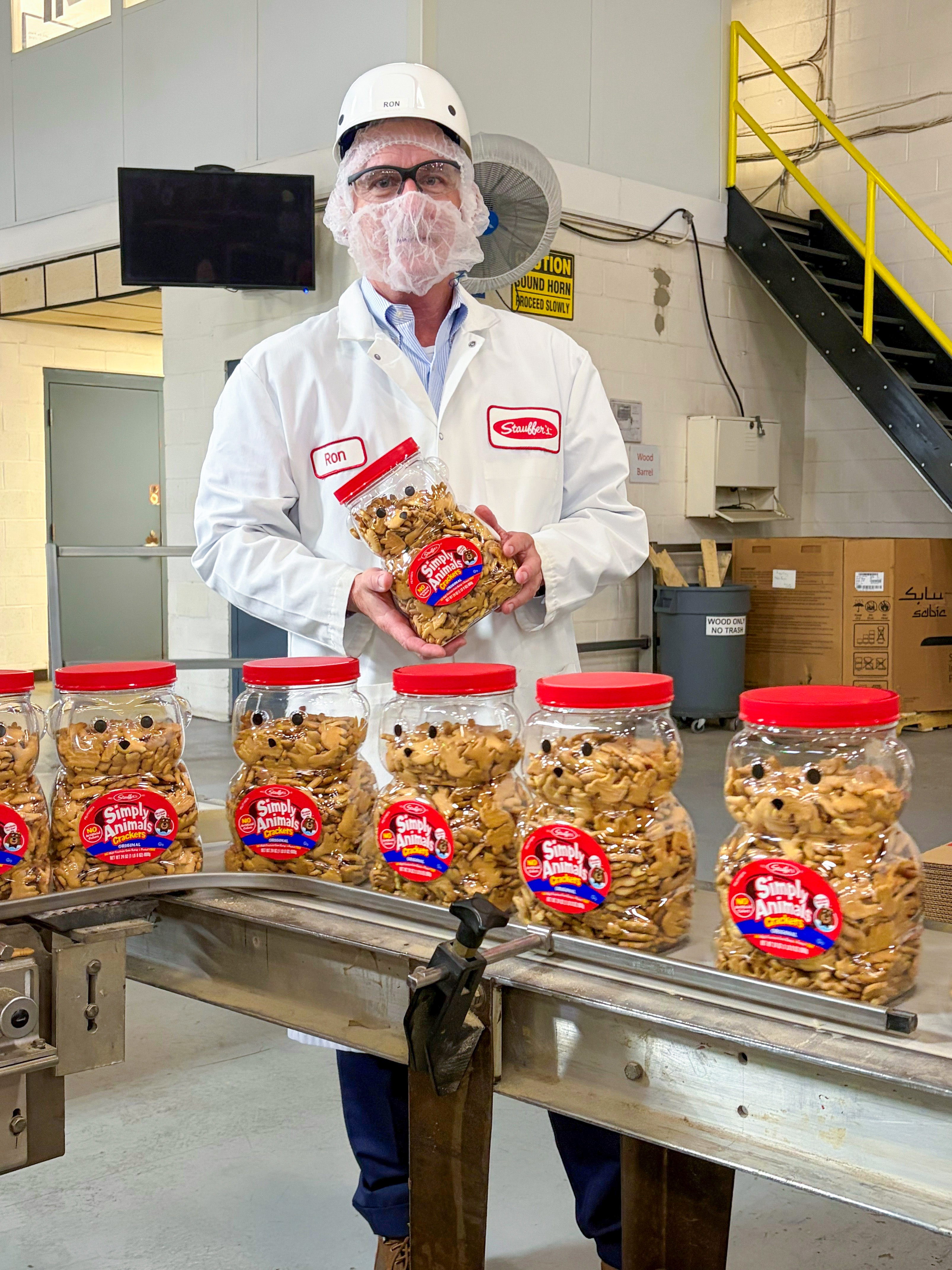 A man wearing a hard hat and hair net holds a container of animal crackers near a manufacturing line in a food processing plant.