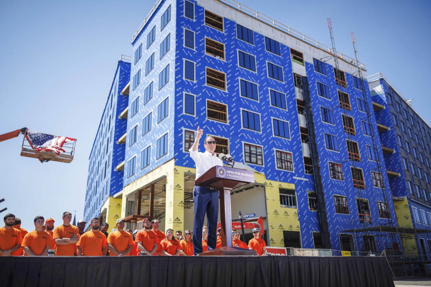 Governor Josh Shapiro waves at a crowd from a podium. Behind him is an under-construction apartment building and a large group of construction workers wearing orange.