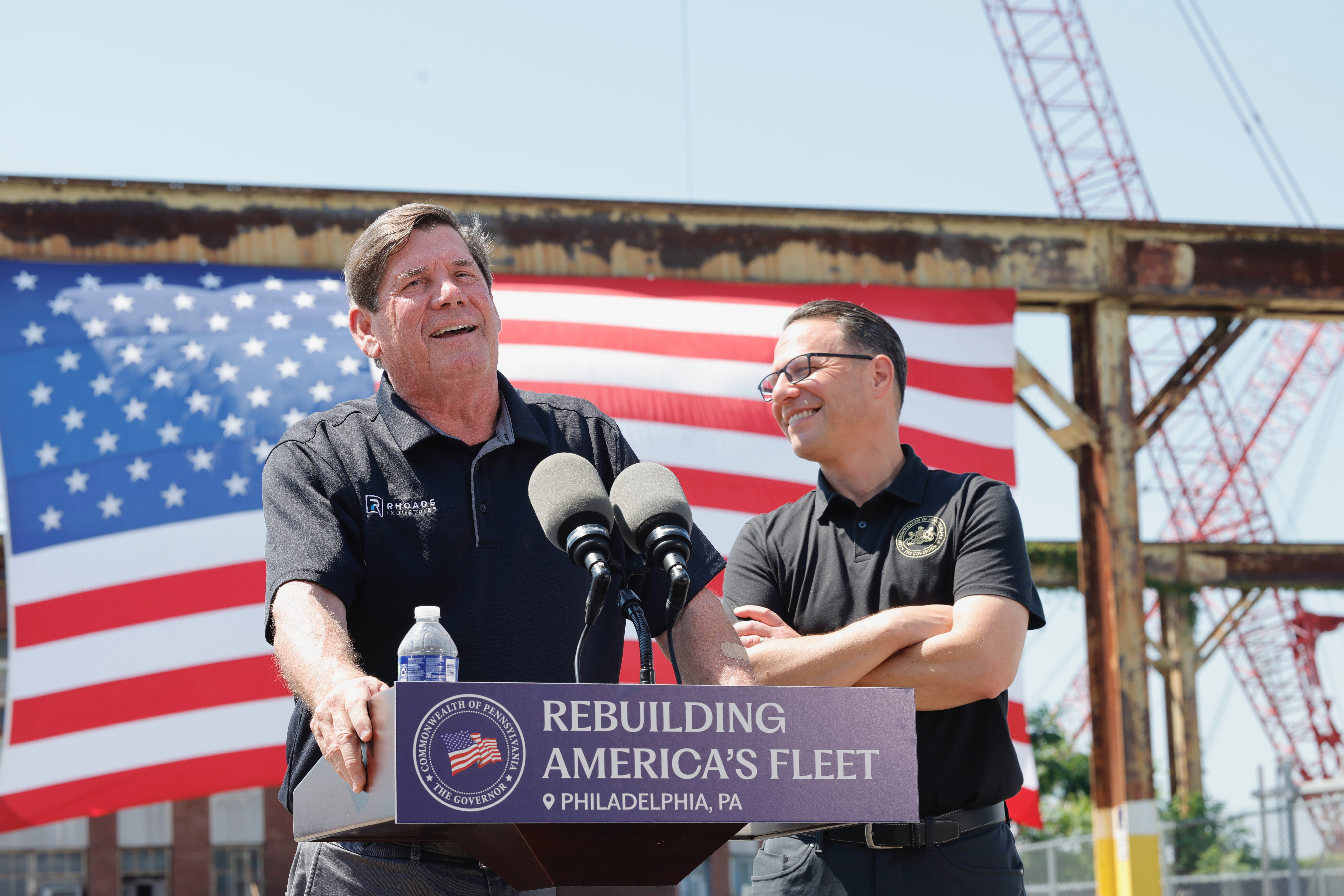 CEO of Rhoads Industries speaks into a microphone at a podium, with Gov. Josh Shapiro standing by his side and smiling. A large American flag is in the background.