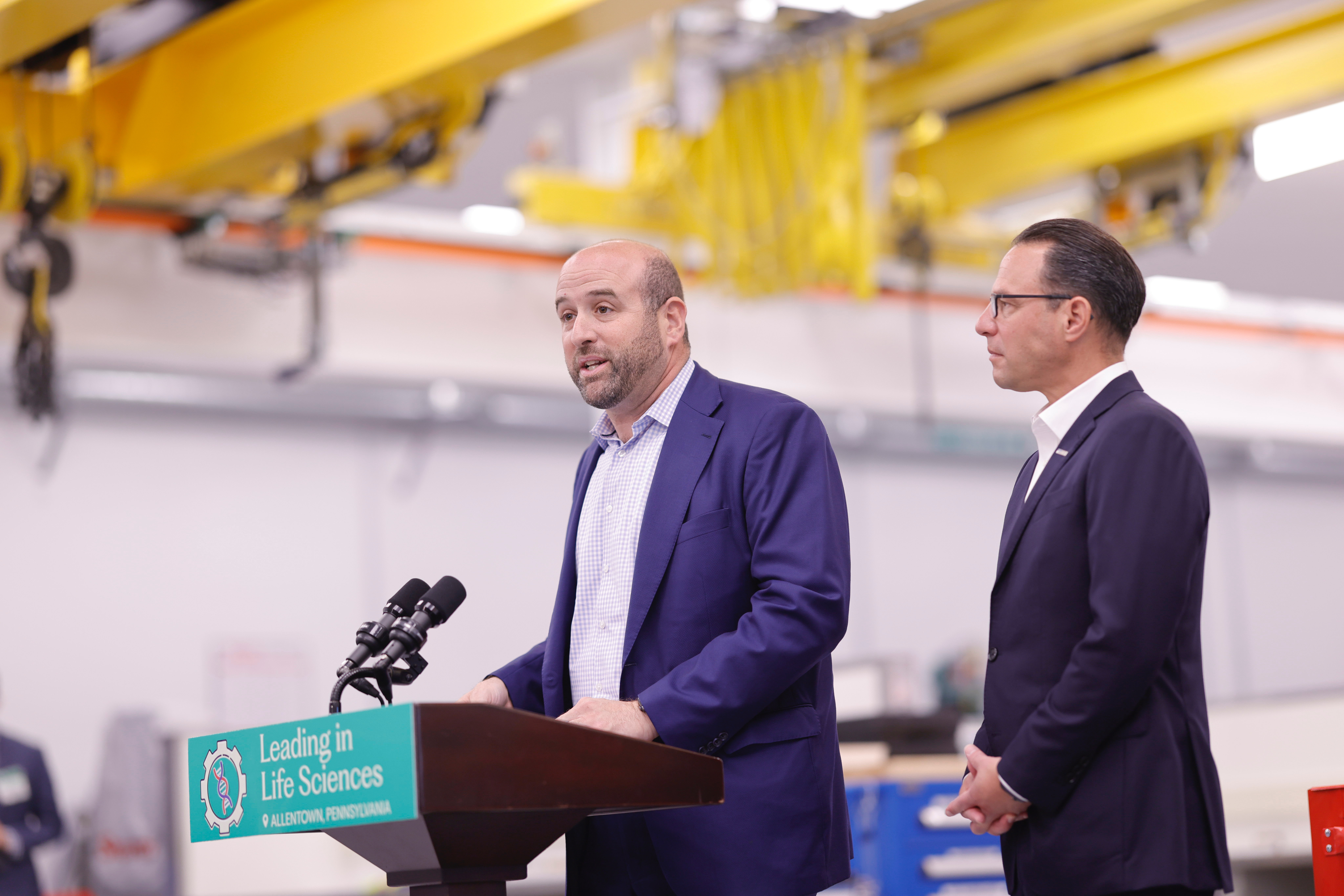DCED Secretary Rick Siger speaks at a podium in an industrial warehouse with Governor Josh Shapiro standing beside him