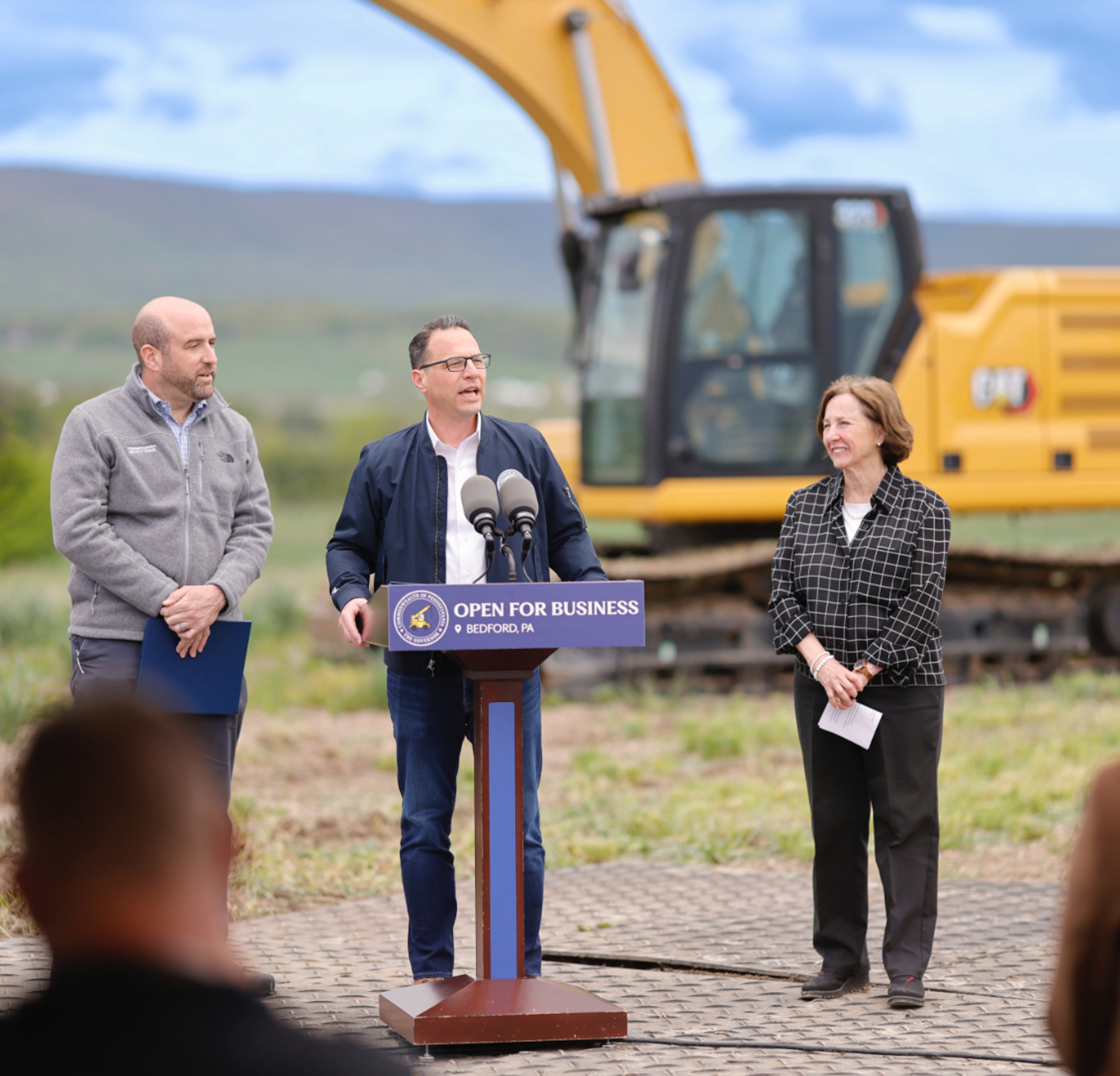 Pennsylvania Governor Josh Shapiro speaks at a podium at a construction site. An excavator and rolling hills are visible in the background. Standing on either side of him are Pennsylvania's Secretary Rick Siger, and Bette Slayton of the Bedford County Development Association