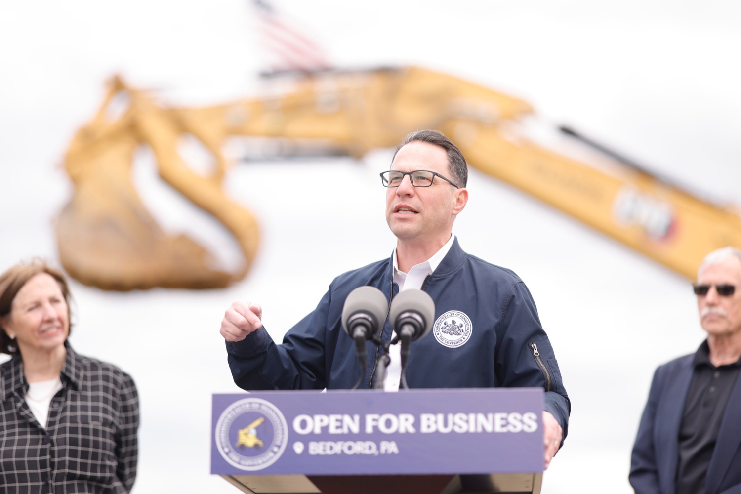 Gov. Josh Shapiro speaks a podium at a construction site. The arm of an excavator is visible in the background.
