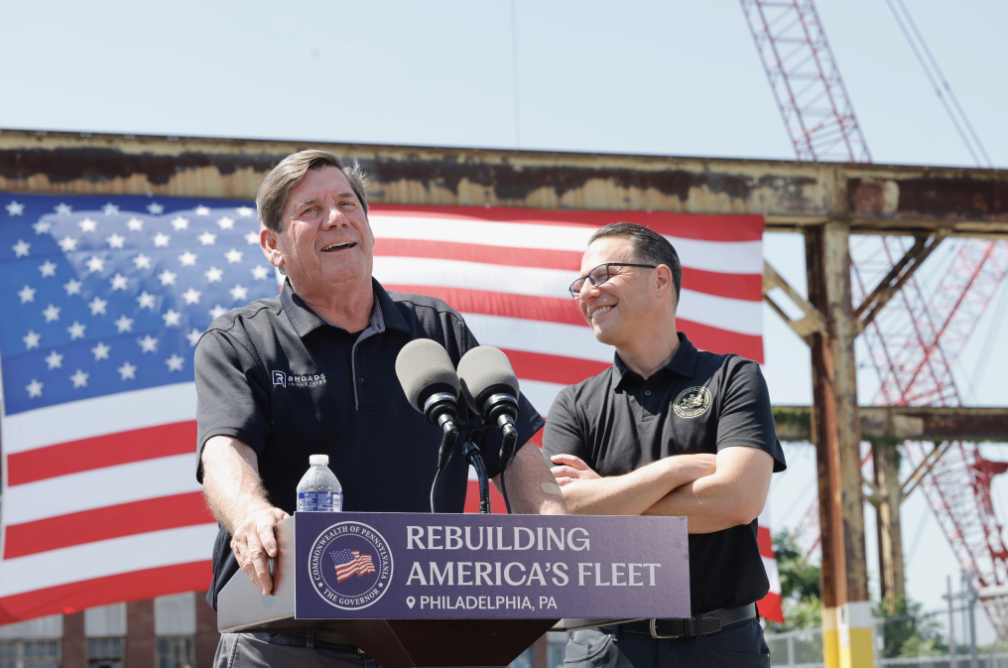 CEO of Rhoads Industries speaks into a microphone at a podium, with Gov. Josh Shapiro standing by his side and smiling. A large American flag is in the background.