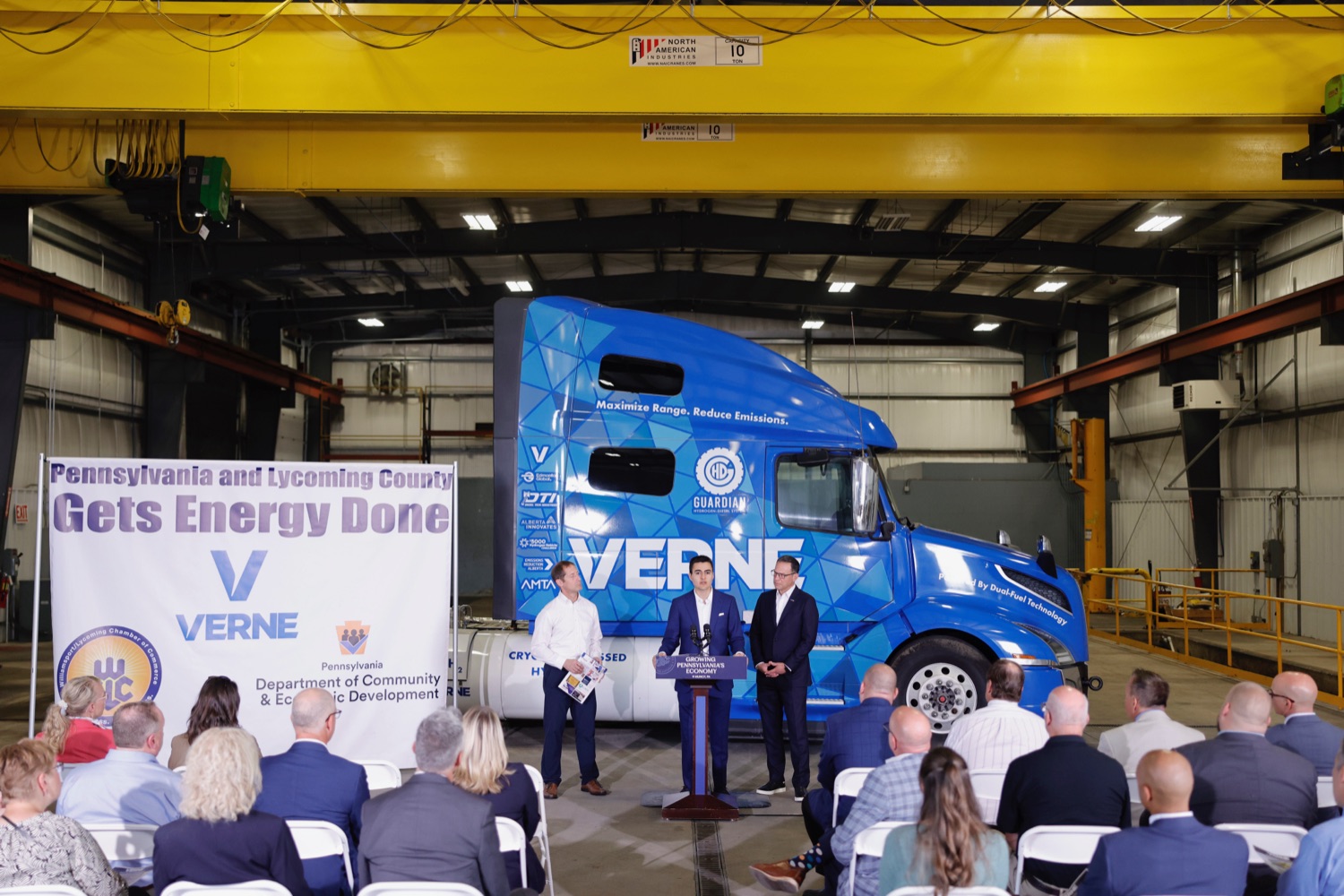 Governor Josh Shapiro and two businesspeople speak at a podium in front of a semitruck cab. A crowd watches. To the left of the podium is a banner that reads "Pennsylvania and Lycoming County Gets Energy Done"