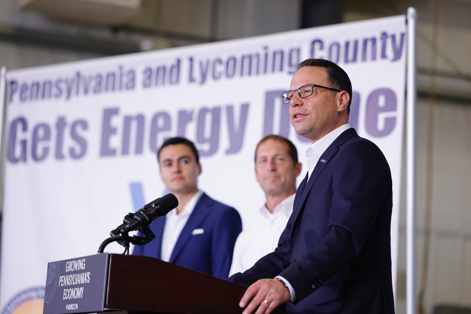 Governor Josh Shapiro speaks at a podium at a news event. Behind him are two corporate officials and a banner that reads "Pennsylvania and Lycoming County Gets Energy Done"