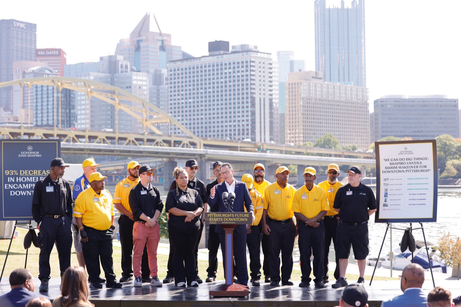 Governor Josh Shapiro speaks at a podium outdoors with a large crowd of construction workers standing behind him. The Pittsburgh skyline is visible in the background..