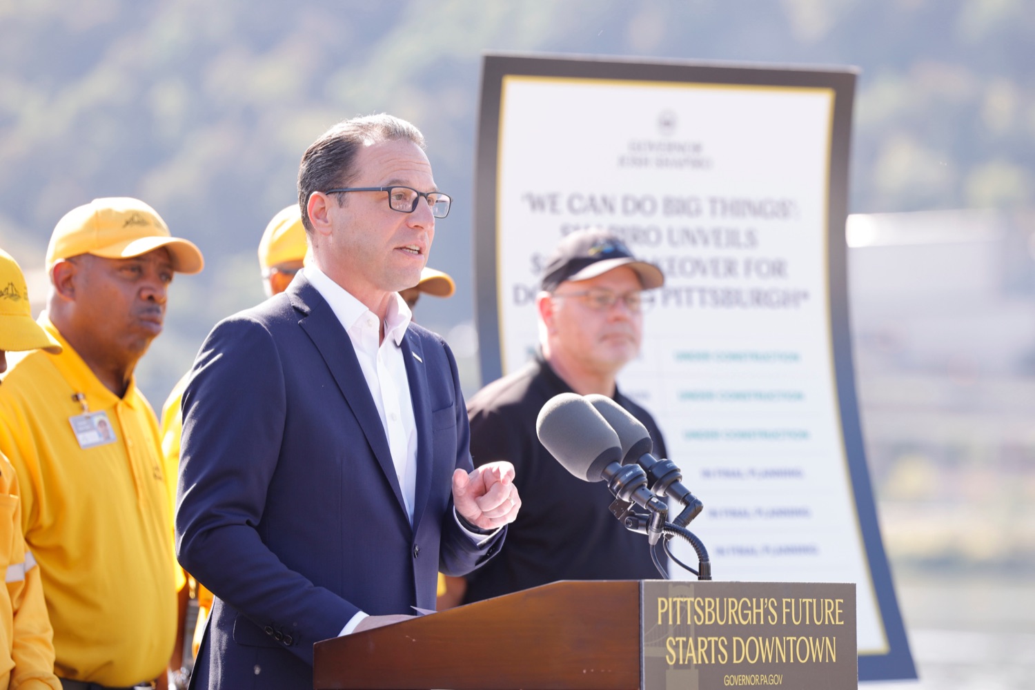Governor Josh Shapiro speaks at a podium outdoors with a small crowd of construction workers standing behind him,