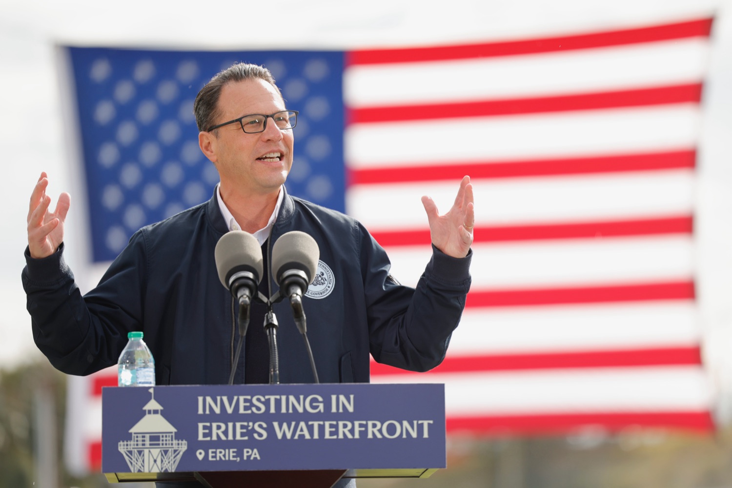 Governor Josh Shapiro speaks at a podium outdoors in front of a large American flag
