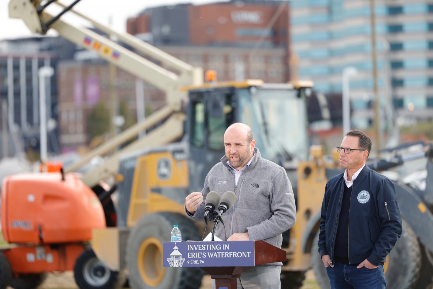DCED Secretary Rick Siger speaks at a podium outdoors with construction equipment in the background. Governor Josh Shapiro stands next to him.