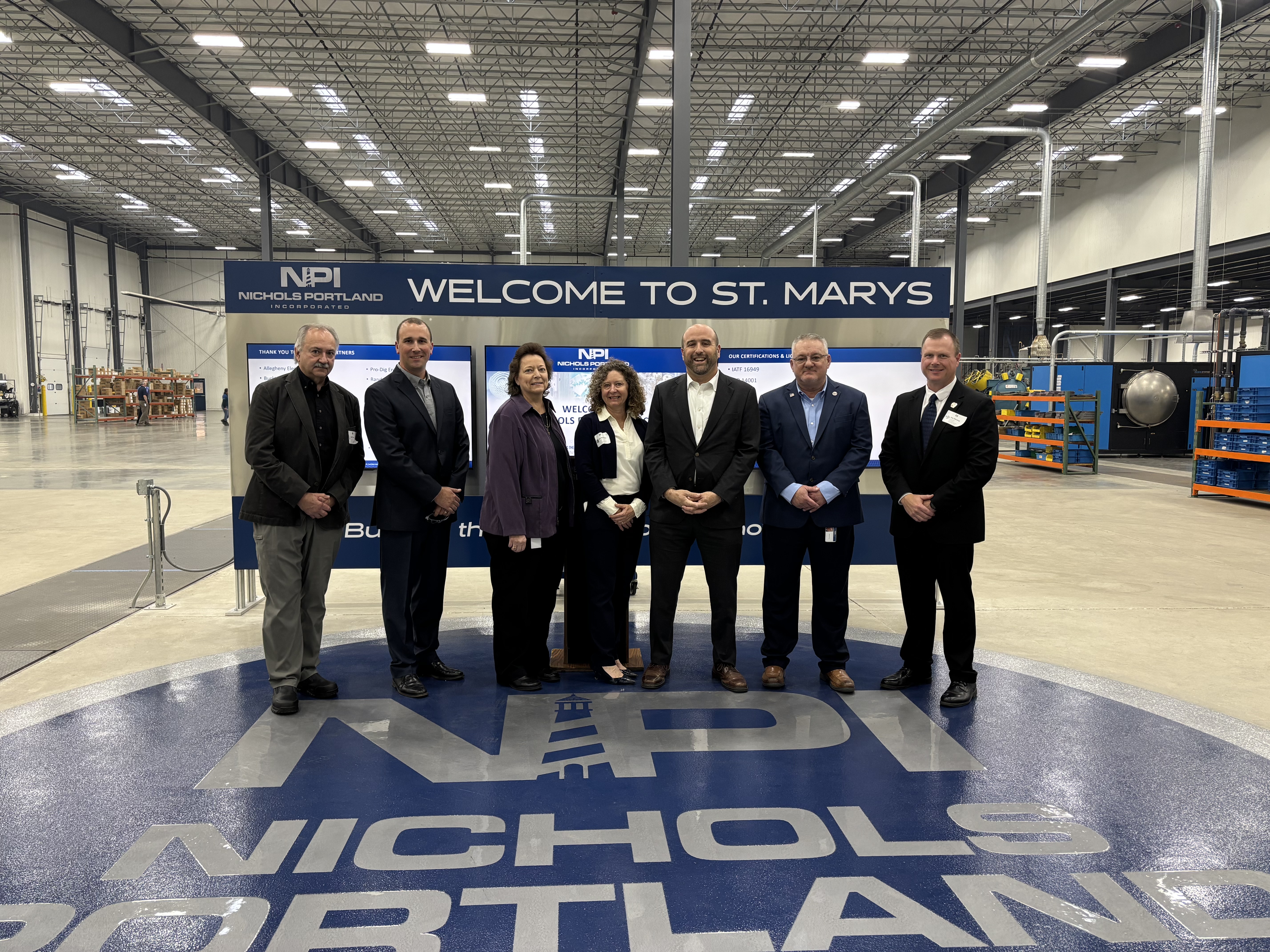 Secretary Rick Siger and a group of executives and officials pose for a group photo in a large fabrication warehouse.