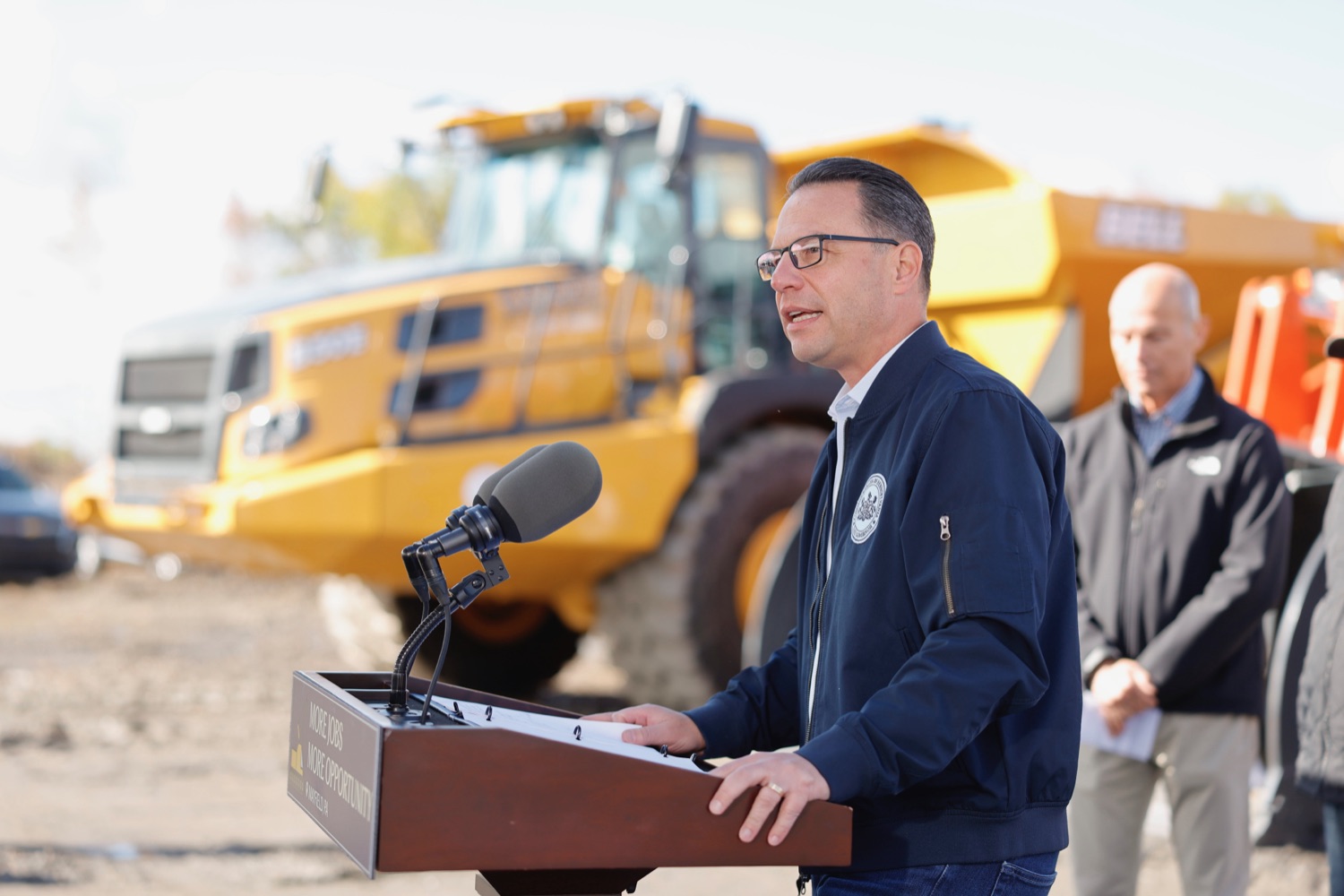 Governor Shapiro speaks at a podium at a construction site. A large dump truck is visible in the background.