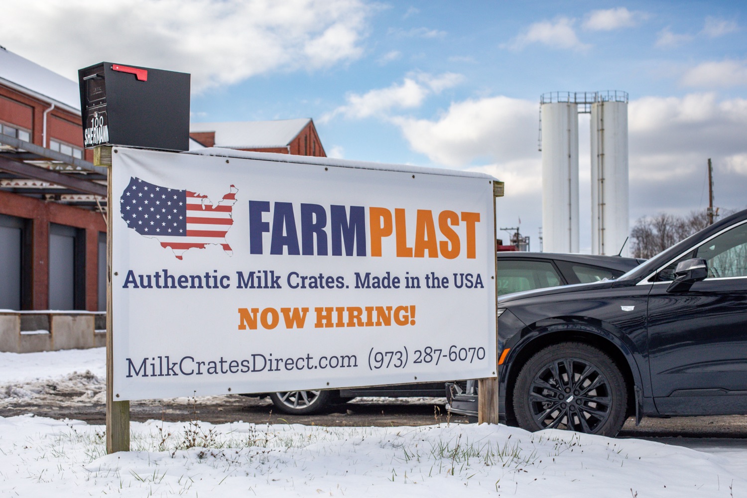 A sign in the parking lot outside a manufacturing facility on a snow day. The sign reads "Farm Plast. Authentic Milk Crates, Made in the USA. Now Hiring!"