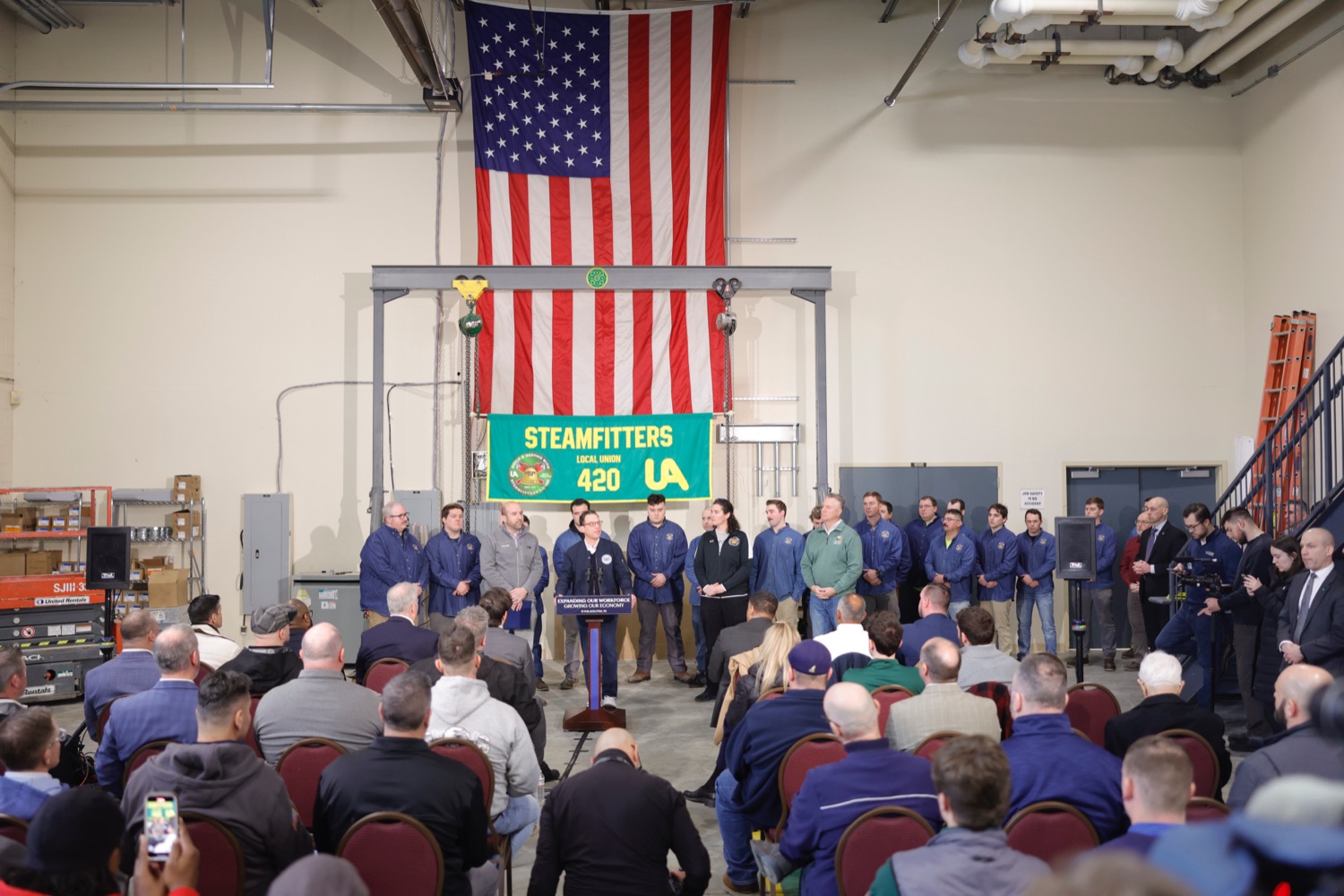 A large crowd watches Governor Josh Shapiro speak at a podium in an industrial building. A large American flag hangs on the wall behind him.