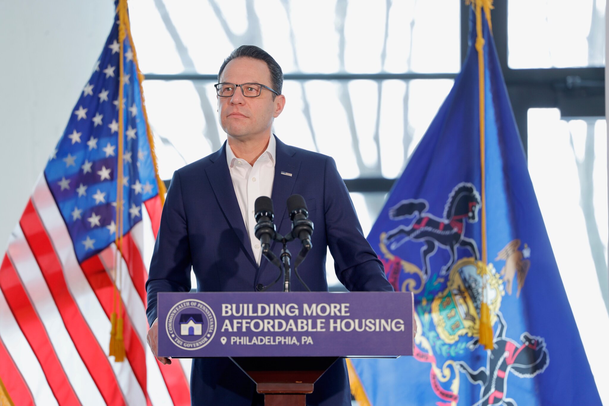 Governor Josh Shapiro speaks at a podium indoors. The US and Pennsylvania flags stand behind him.
