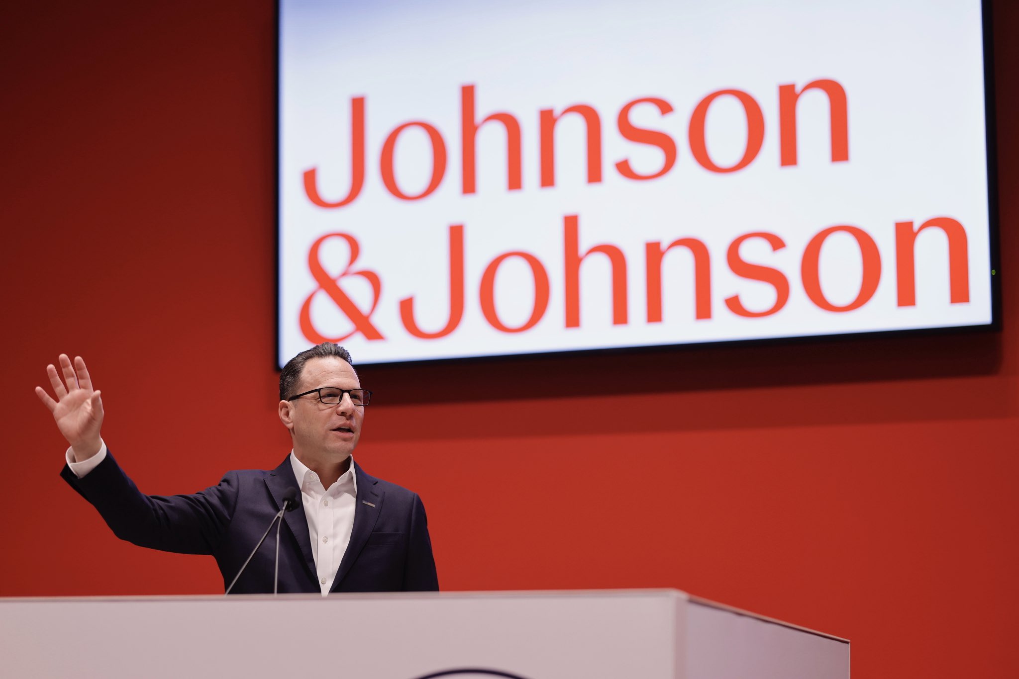 Pennsylvania Governor Josh Shapiro speaks at a podium. The Johnson & Johnson company logo is projected on the wall behind him.