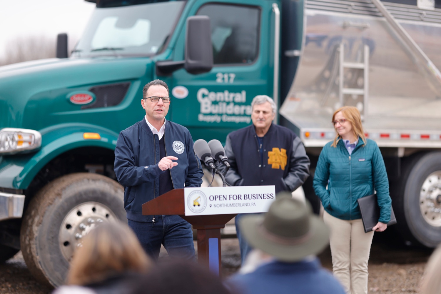 Governor Josh Shapiro speaks at a podium at a construction site. A large truck is parked behind him.
