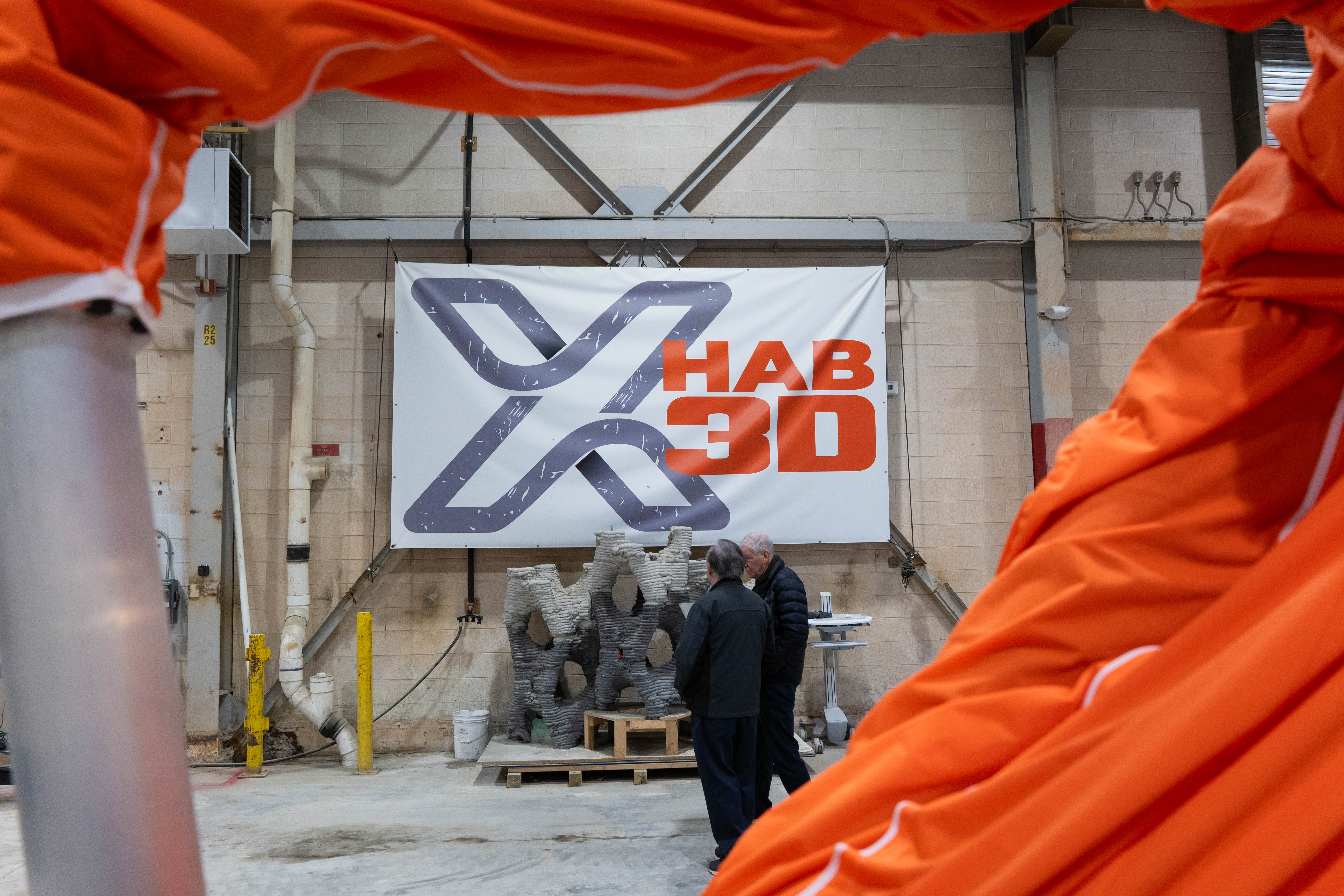A sign for the company X-Hab 32 hangs on the wall in an industrial building. Two men converse in front of it.