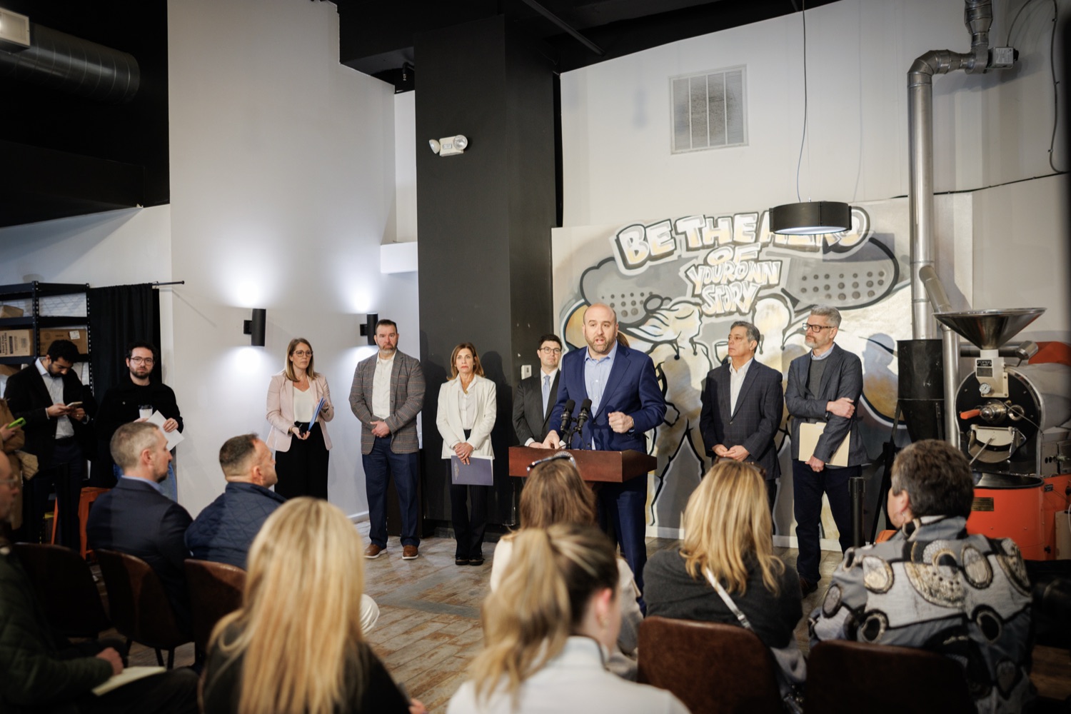 DCED Secretary Rick Siger speaks at a coffee shop at a podium. A crowd of professionals stands behind him.