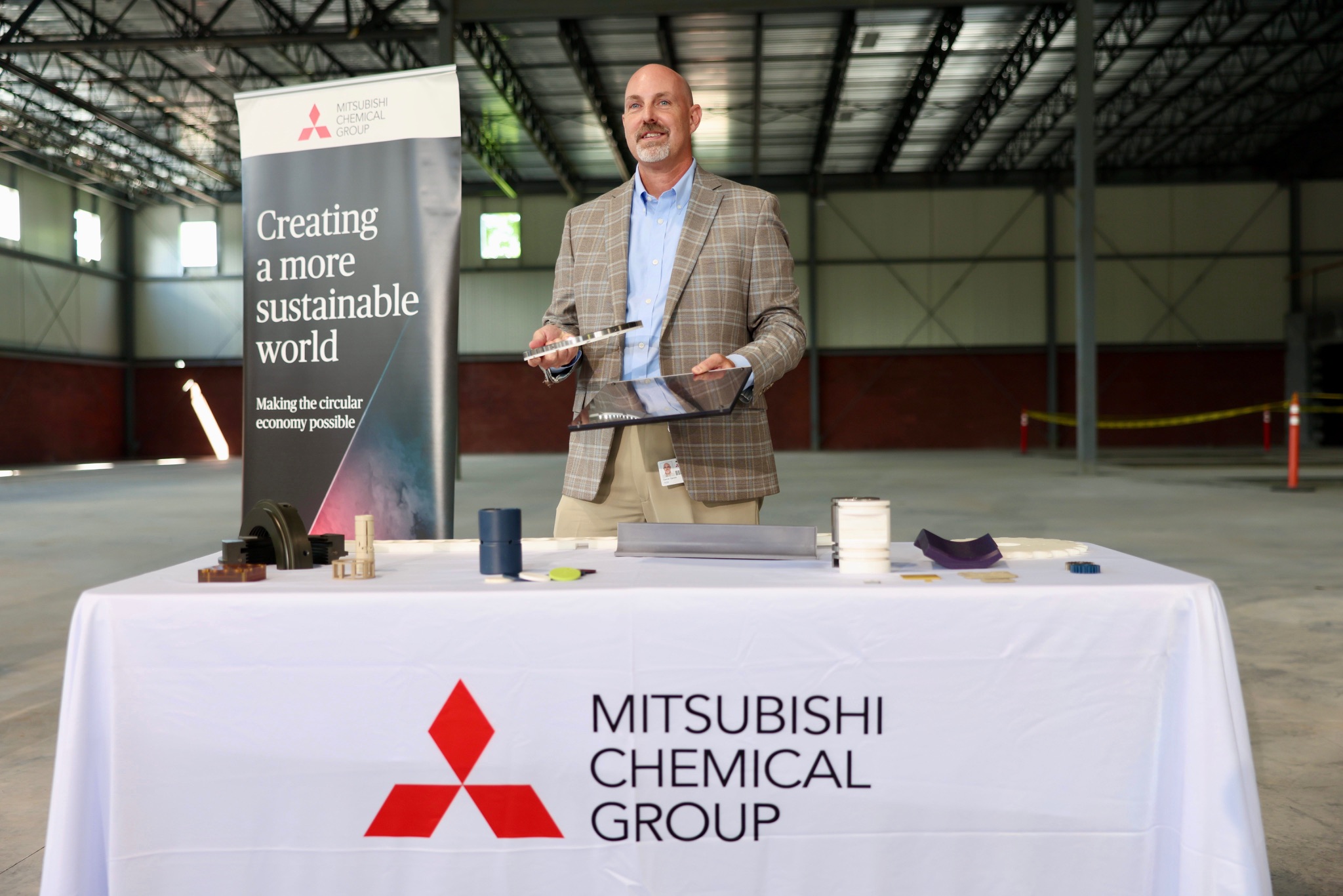 A man stands at an informational booth branded with the logo of Mitsubishi Chemical Group. He is in a large industrial warehouse.