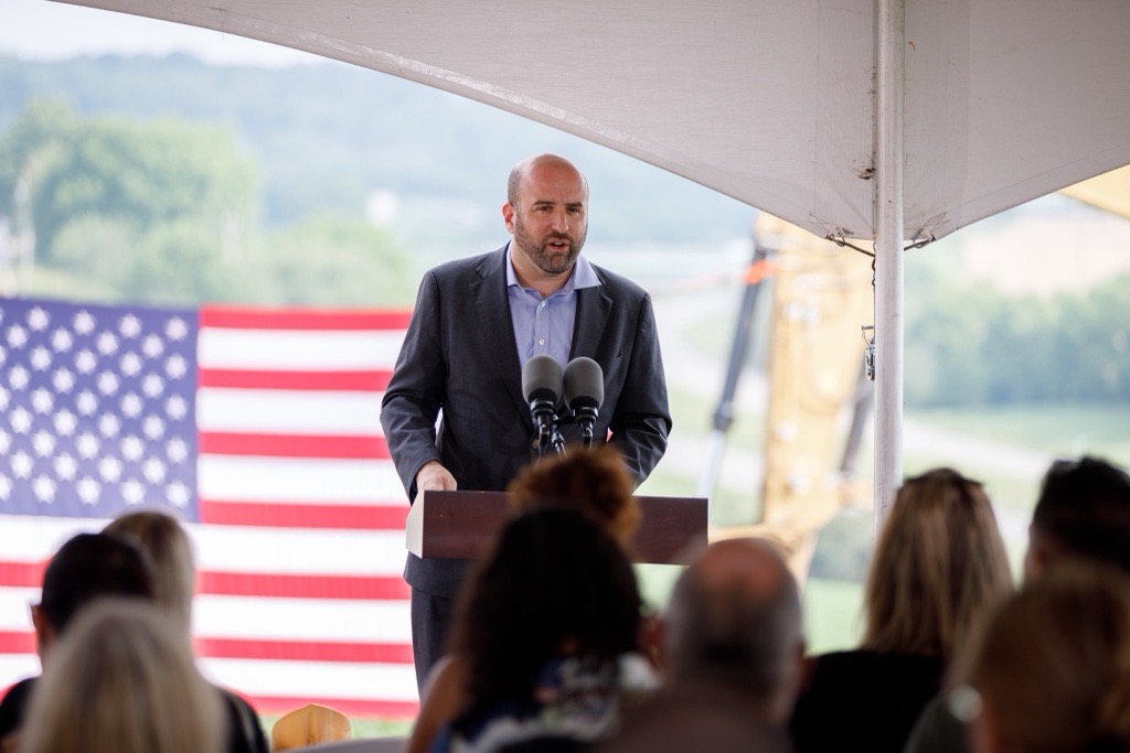 DCED Secretary Rick Siger speaks at a podium at a construction site. A large American flag is visible in the background.