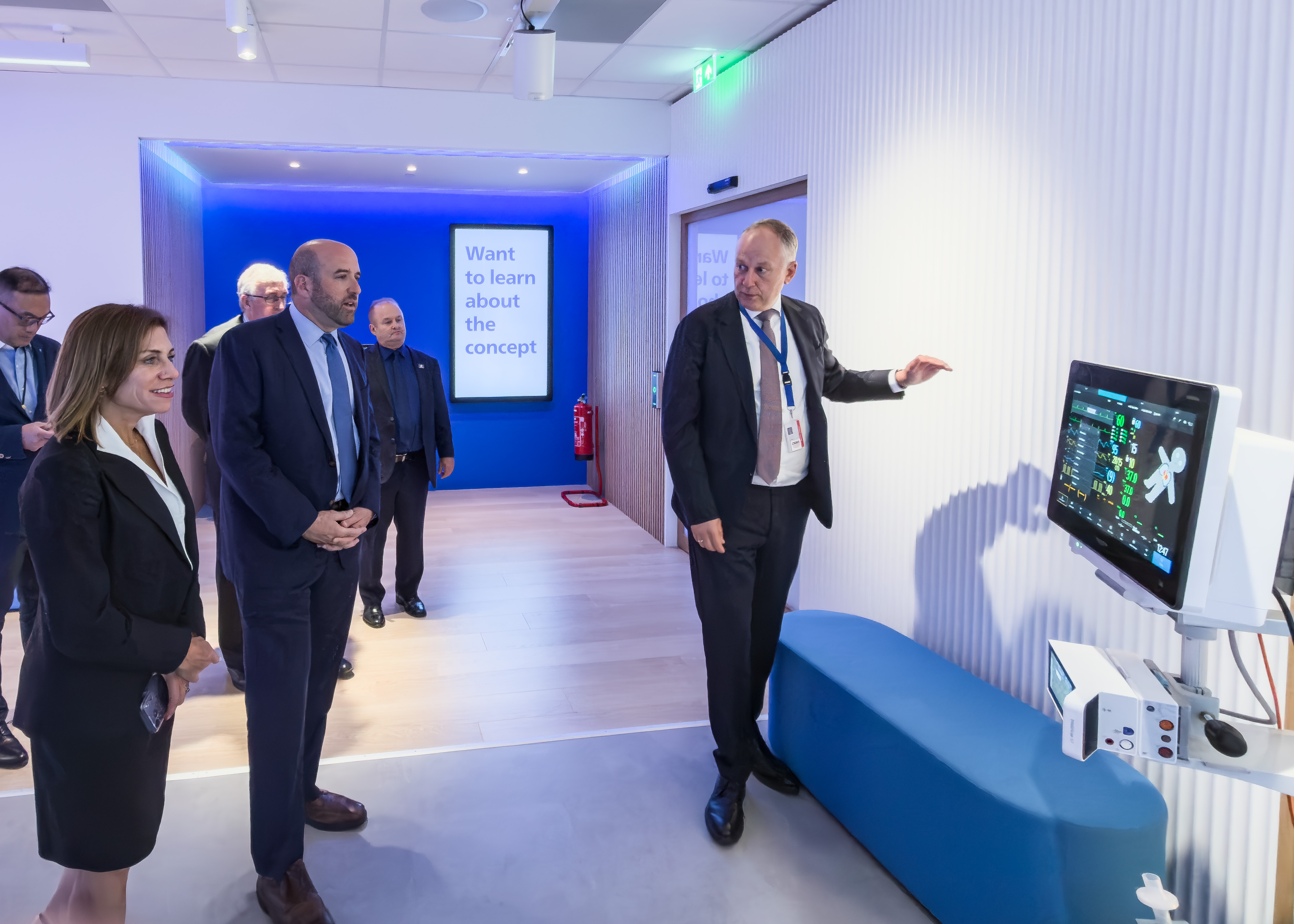 A businessperson from the company Philips takes Secretary Rick Siger and a group of businesspeople on a tour of Philips' facility. The businessperson gestures to an advanced EKG machine.