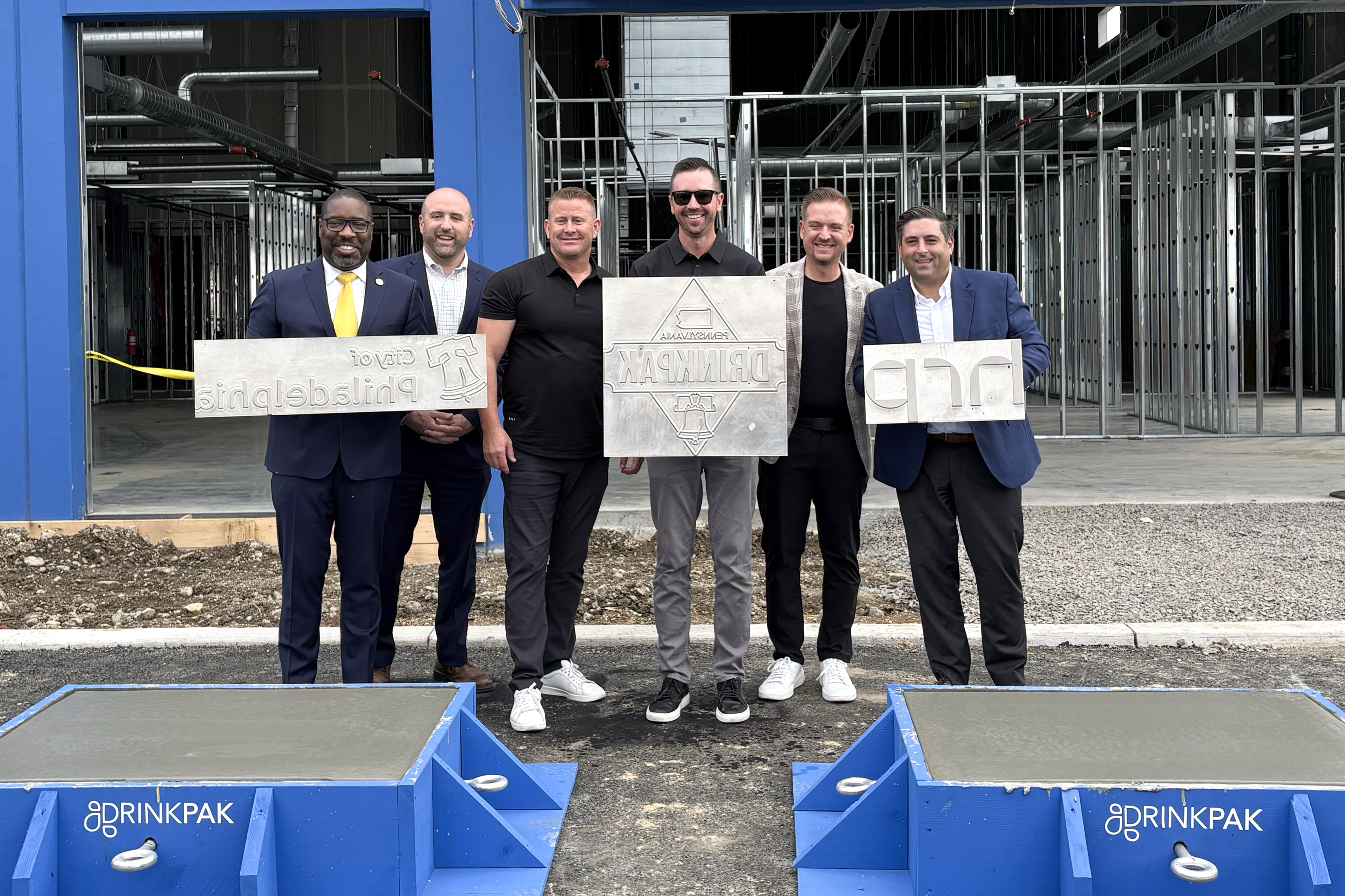 DCED Secretary Rick Siger stands alongside a small group of businesspeople holding silver plaques. A partially constructed building with exposed steel frames is visible behind them.