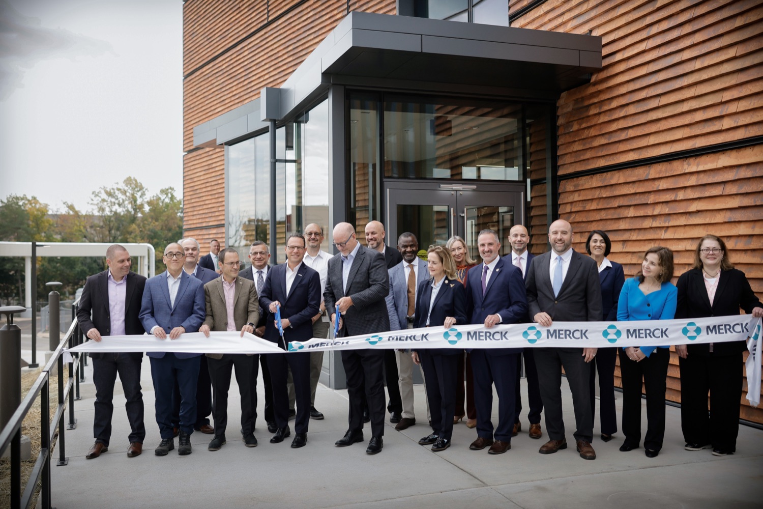 A group of government officials and businesspeople cut a ceremonial ribbon, decorated with the company logo for Merck,  outside a modern office building.
