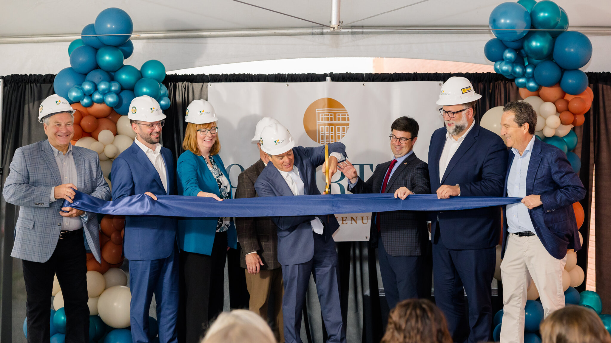 A group of government and business officials cut a ceremonial ribbon while wearing hard hats.