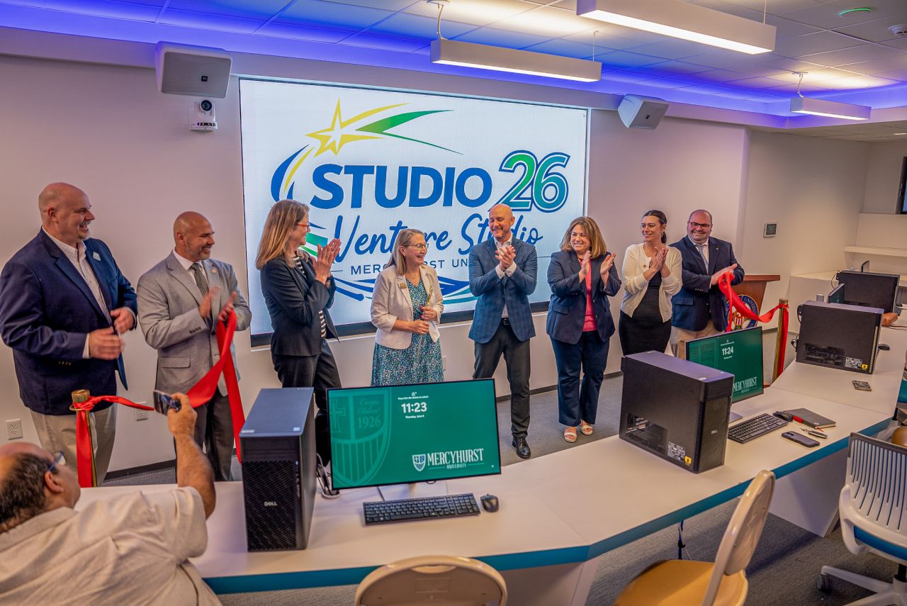 A group of businesspeople, standing in the front of a college classroom, smile and applaud in front of a sign that reads: Studio 26 Venture Studio.