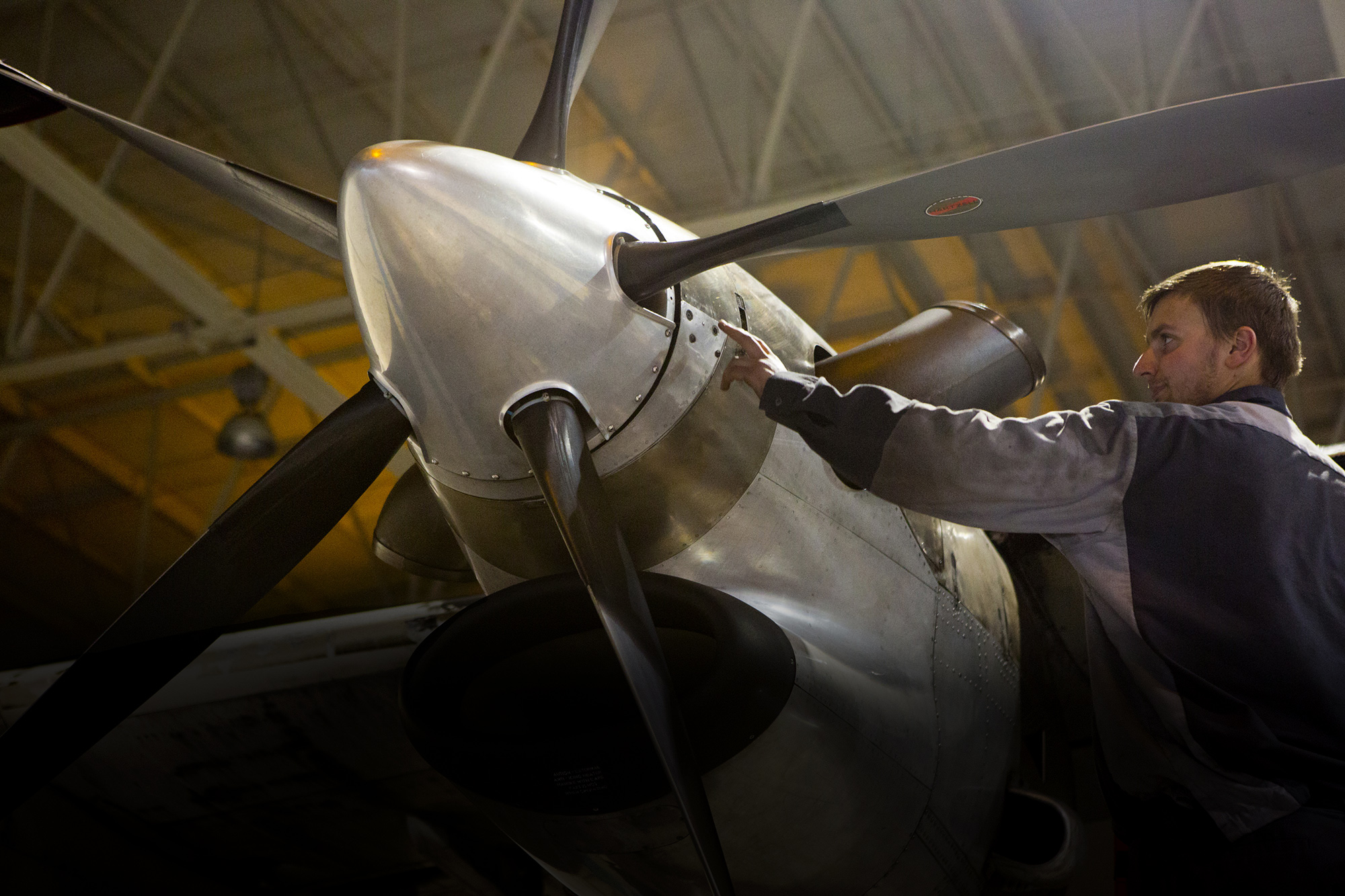 An airplane mechanic repairs a propeller on an aircraft in a dimly lit hangar