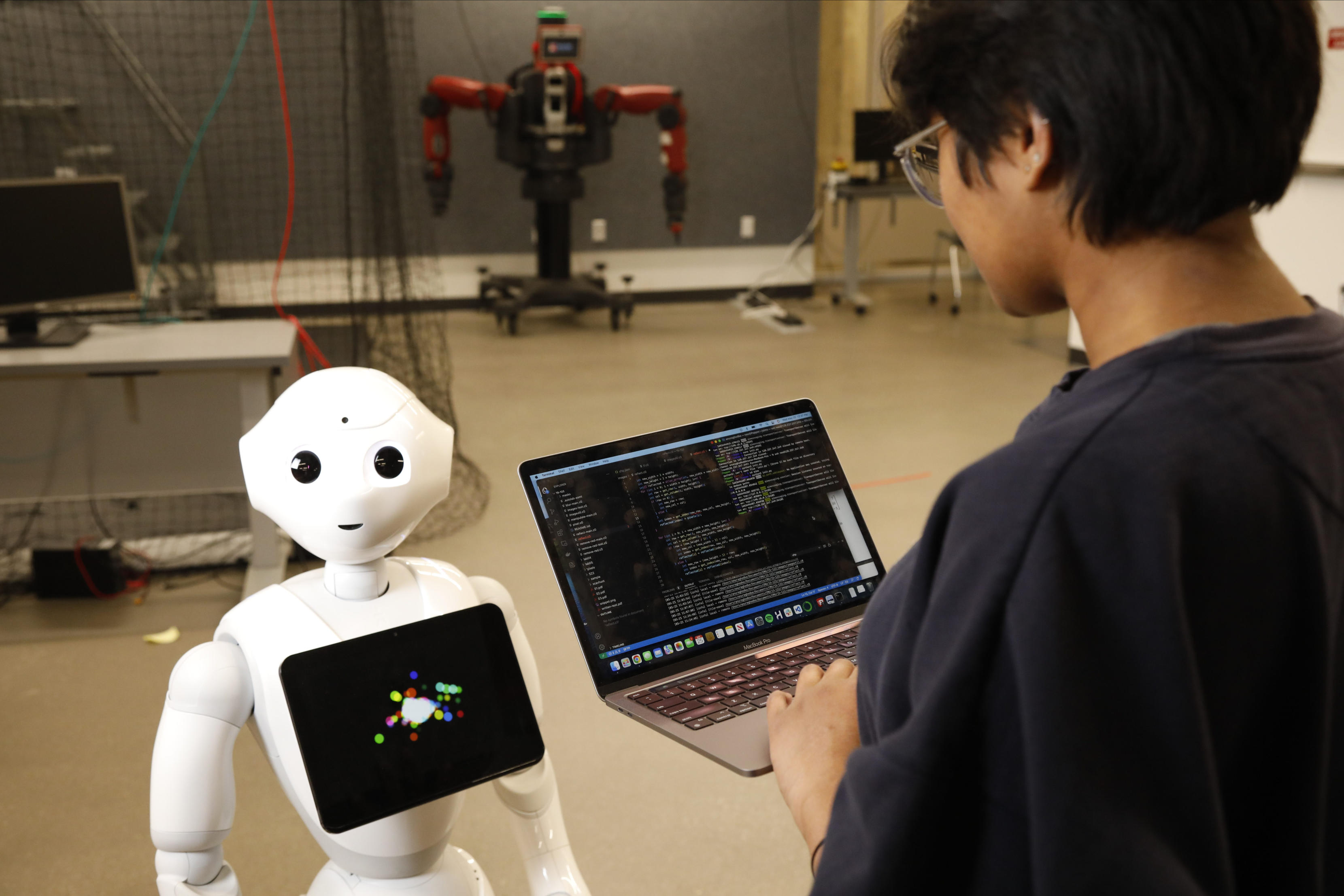 A man holding a laptop controls a humanoid, white plastic robot in a warehouse-like space