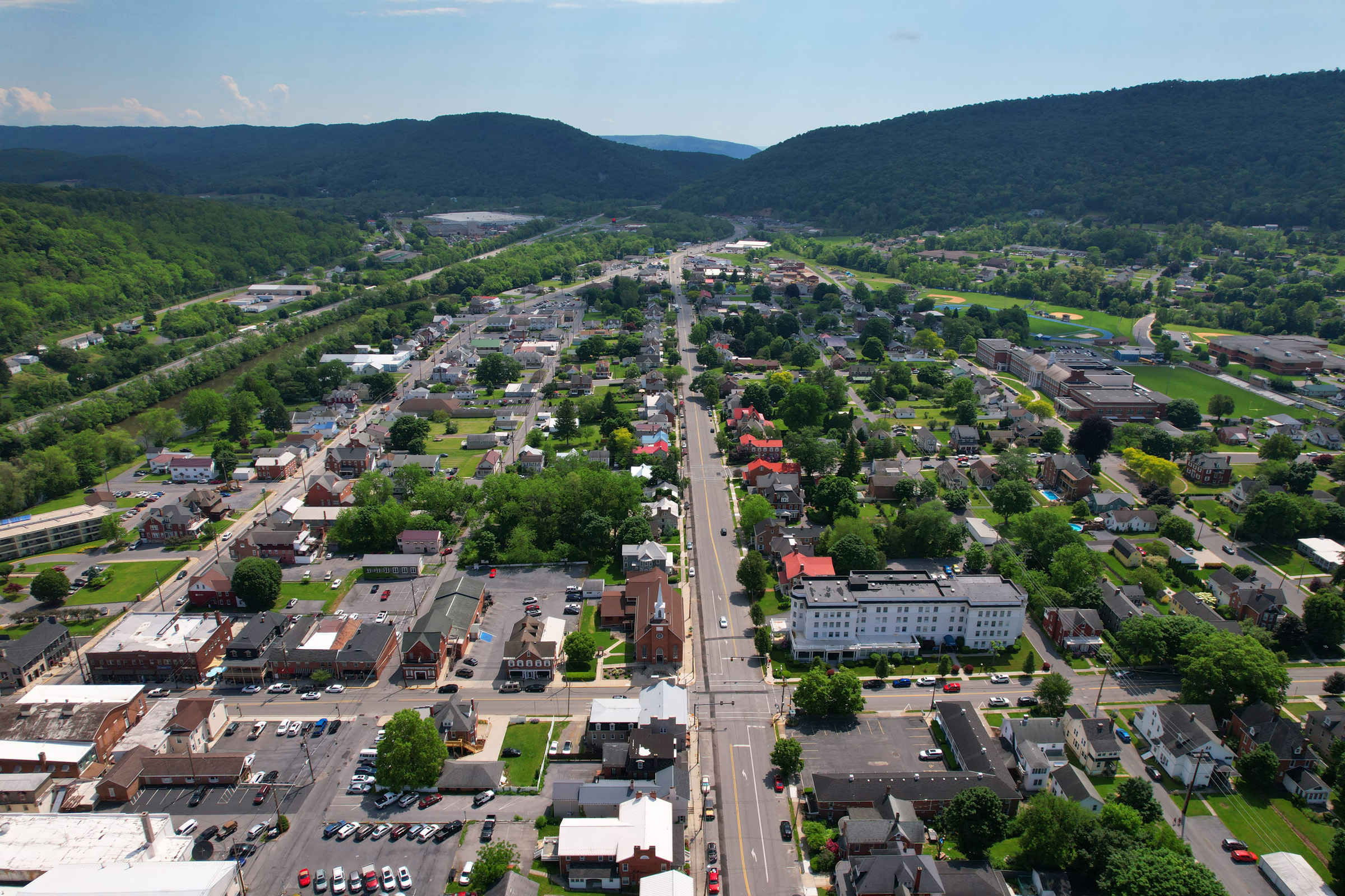 An aerial image of Bedford, PA. Buildings, roads and foliage extend into the distance, surrounded by forest-covered mountains and a blue sky.