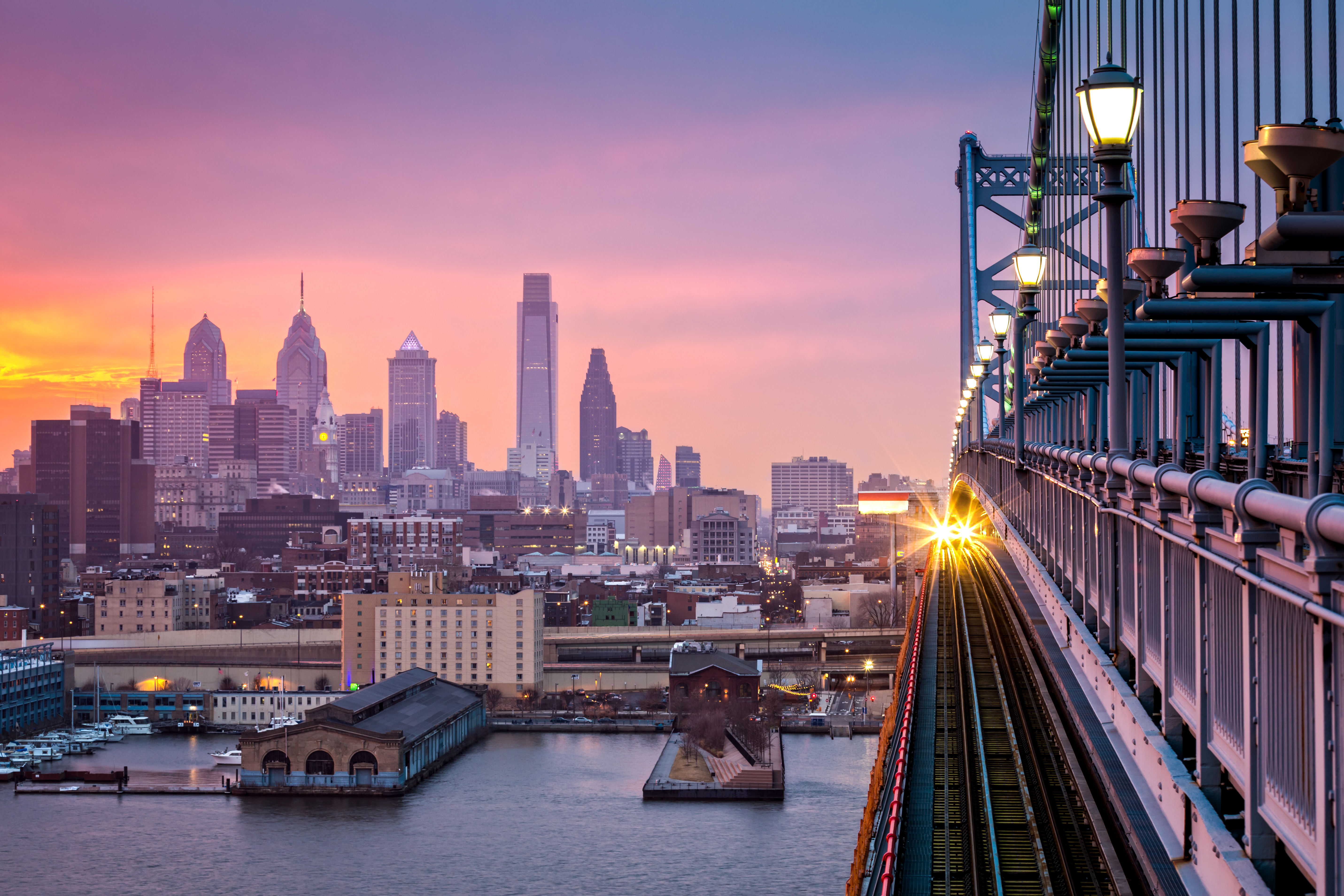 Philadelphia under a hazy purple sunset. An incoming train crosses Ben Franklin Bridge