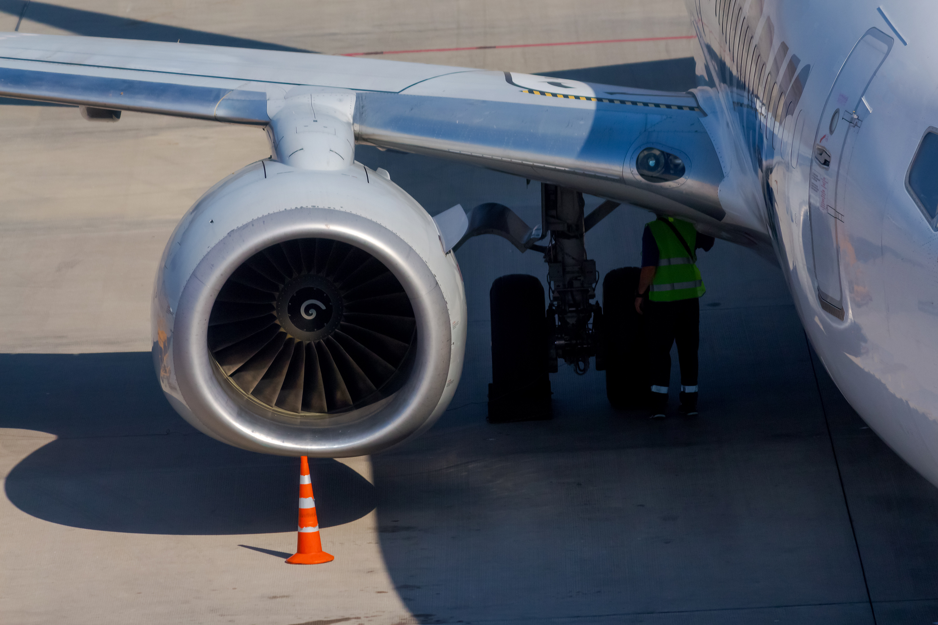 Engine of white commercial airplane docked on apron outside airport terminal. Unrecognisable worker under the wing. Sunny day. Ground service theme.