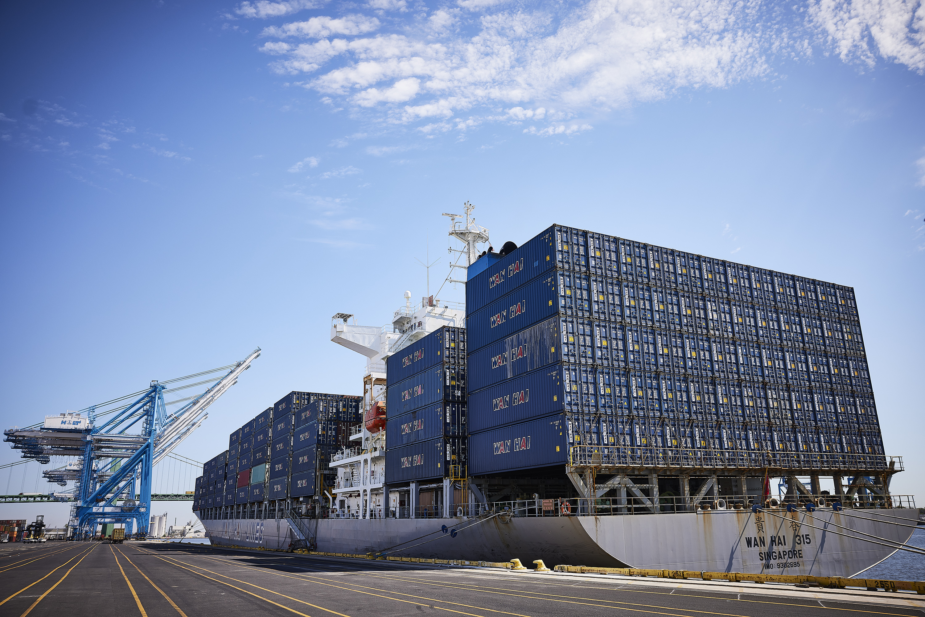 A Wan Hei Lines cargo ship, loaded up with shipping containers, docked at the Port of Philadelphia on a sunny day. Large port cargo cranes are visible in the background.