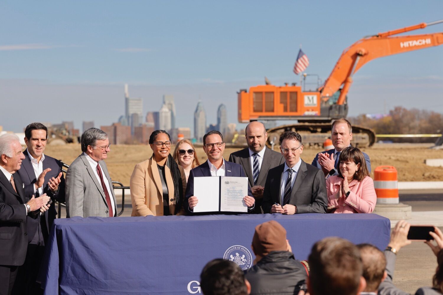 Governor Josh Shapiro, surrounded by a group of government officials and business leaders, smiles and shows an exectutive order document for the just-announced Permit Fast Track program. The Philadelphia skyline and construction equipment are visible in the distance.